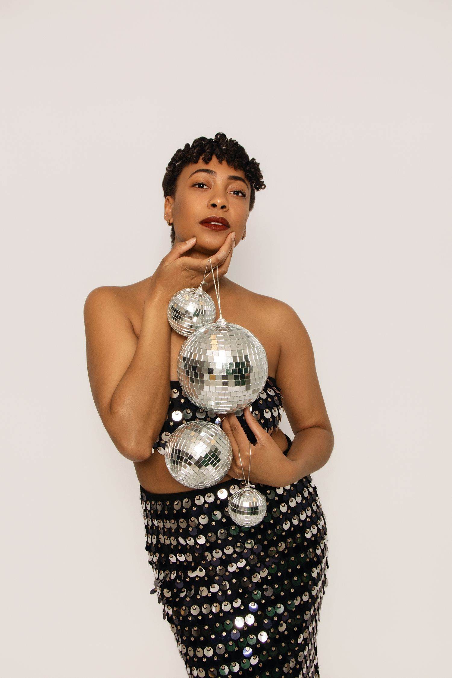 Woman in sequin dress holding silver disco balls poses against white background.