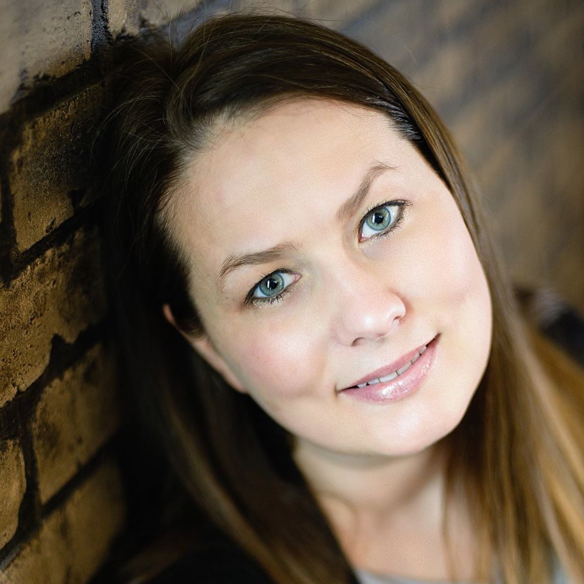 Young woman with blue eyes and brown hair smiling against a brick wall in a professional portrait photo.
