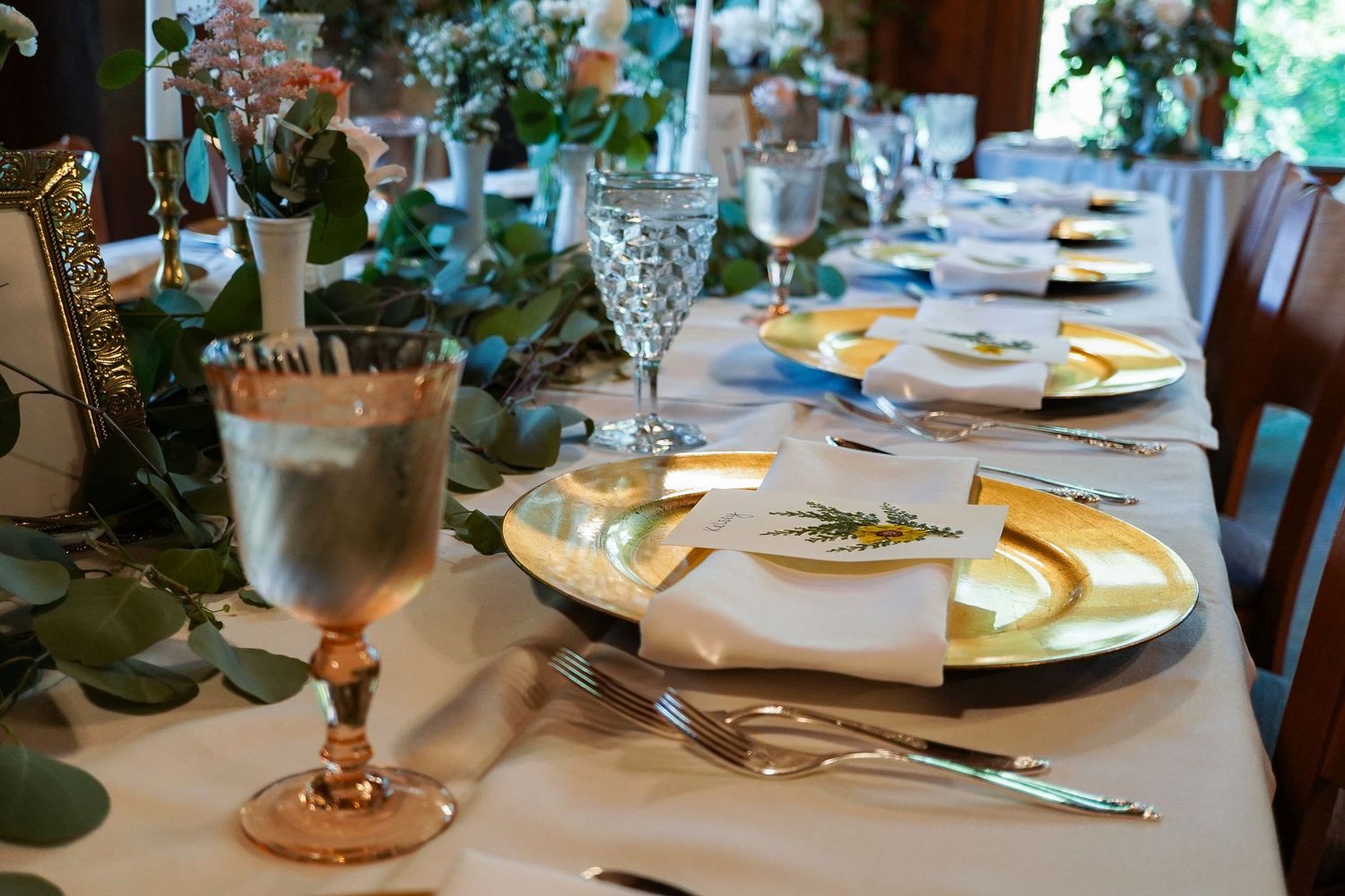 Elegant wedding reception table set with gold chargers, crystal glasses, white napkins, and eucalyptus garland.