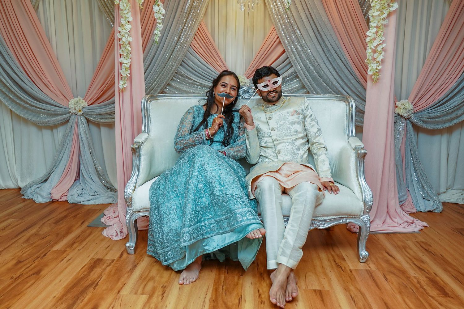 Couple in traditional South Asian wedding attire laughing together on a white sofa with drapes.