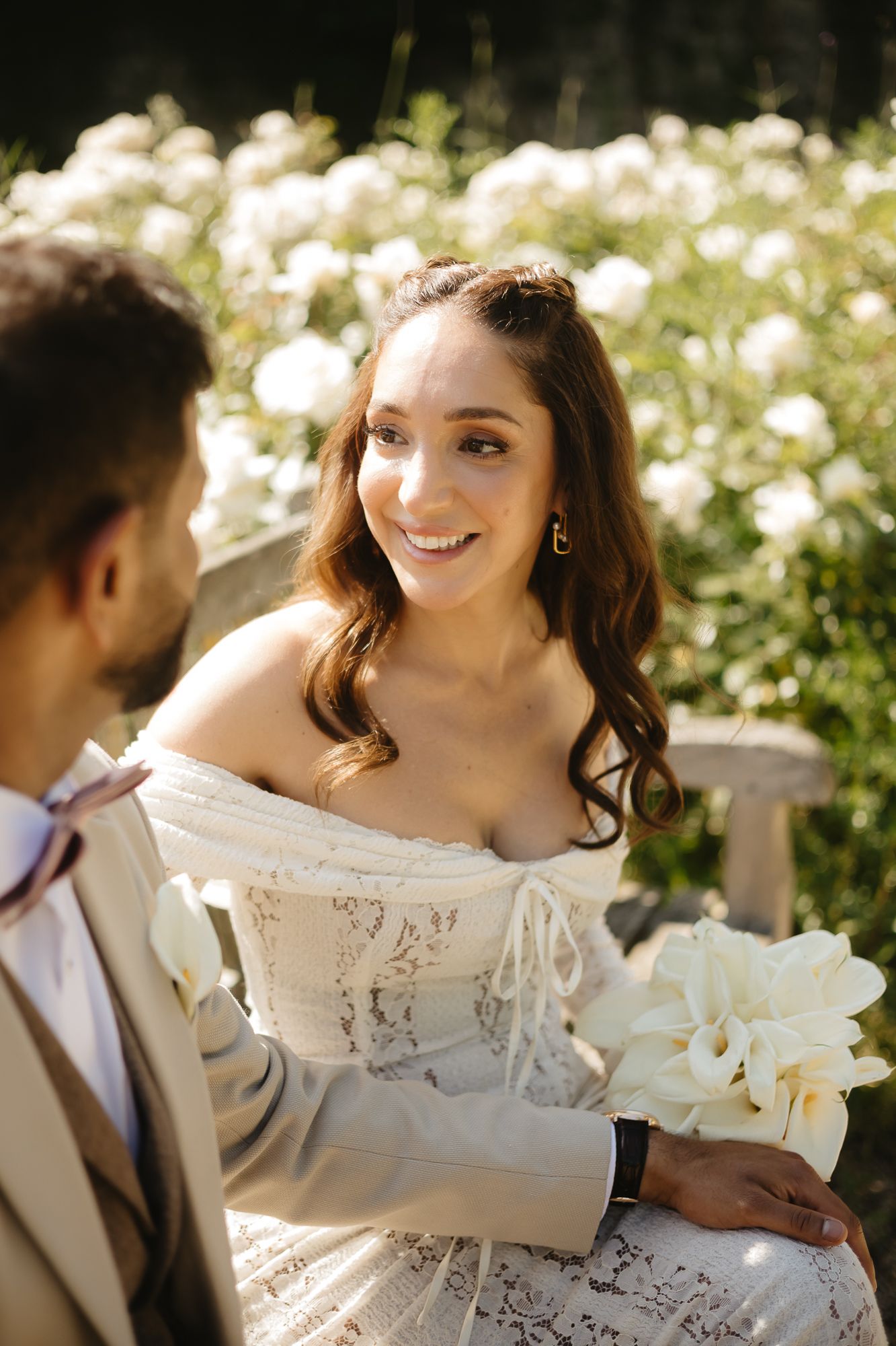 Bride in an off-shoulder lace wedding dress poses in a sunny garden setting.