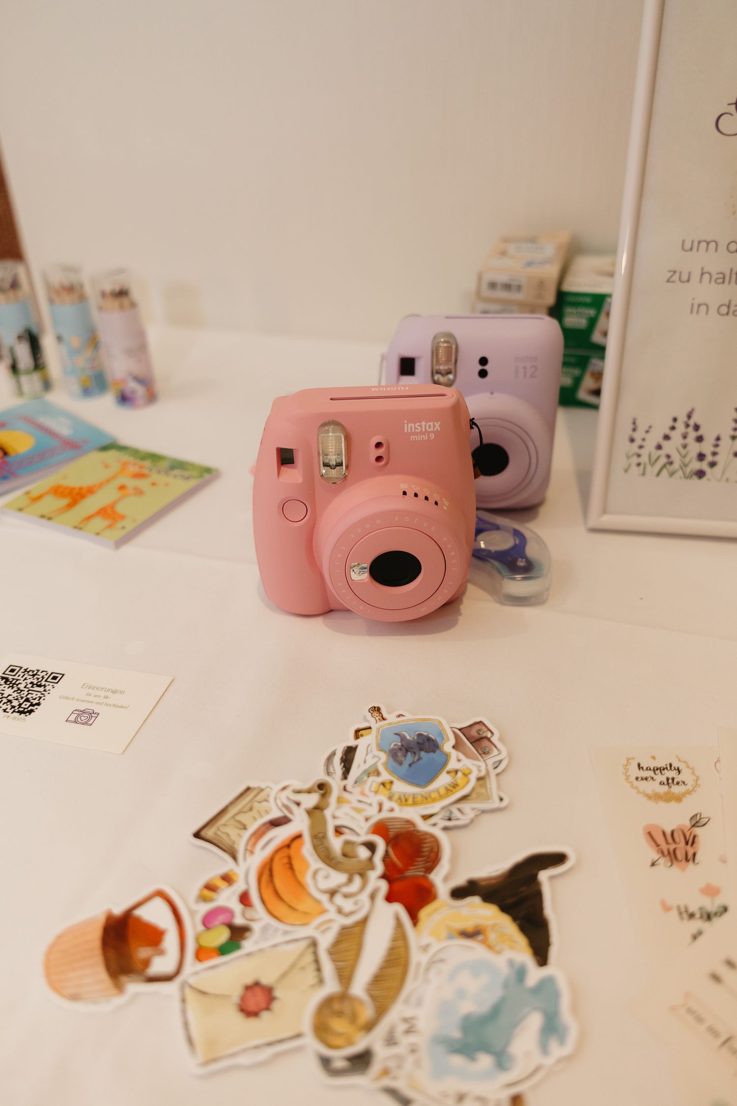 A pink Fujifilm Instax Mini camera sits among scattered stickers and decorative elements on a light surface.