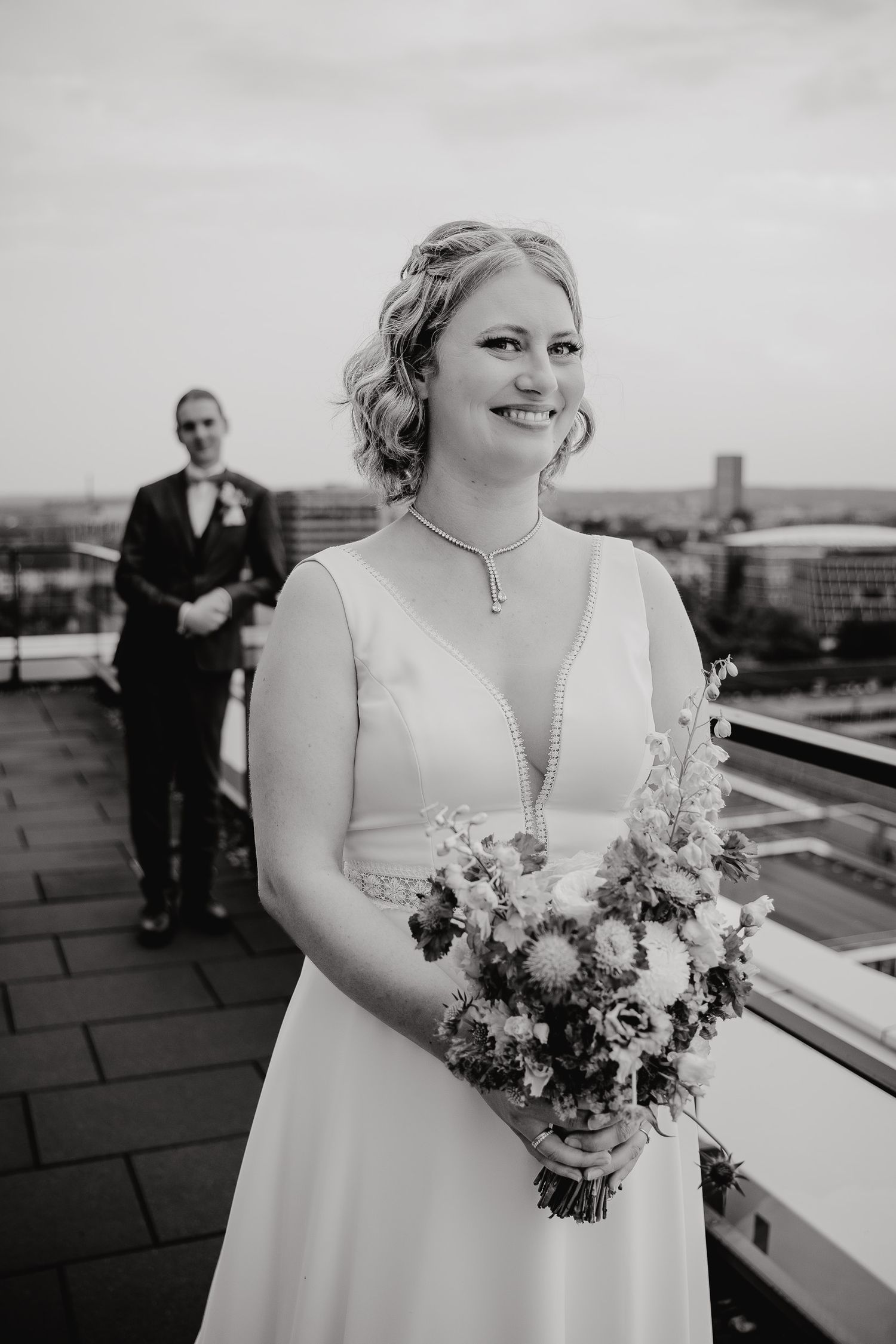 A bride in a white wedding dress holds a floral bouquet on a rooftop terrace with cityscape in background.