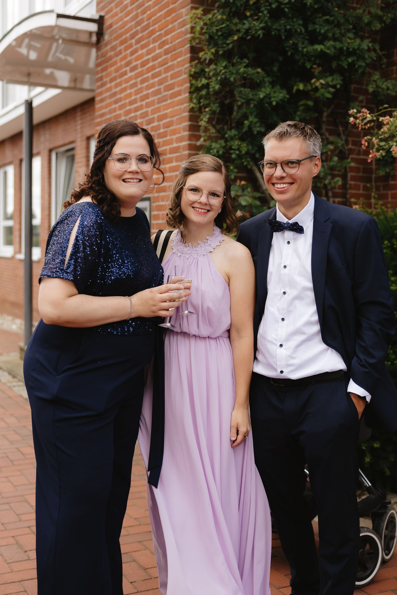 Wedding guests in formal attire gather for photos outside a brick building.
