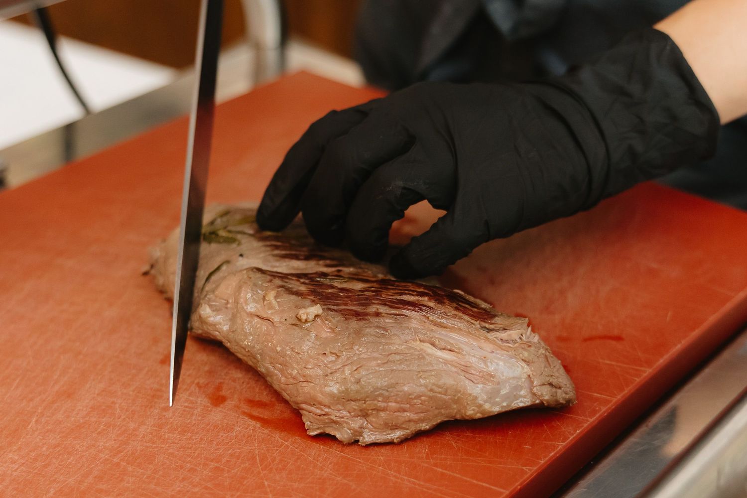 Person wearing black gloves slices raw meat on a red cutting board with a sharp knife.