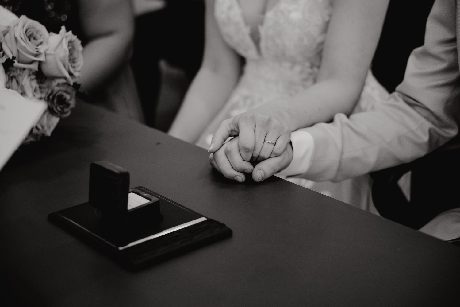 Black and white close-up of newlyweds holding hands at a table during their wedding ceremony.