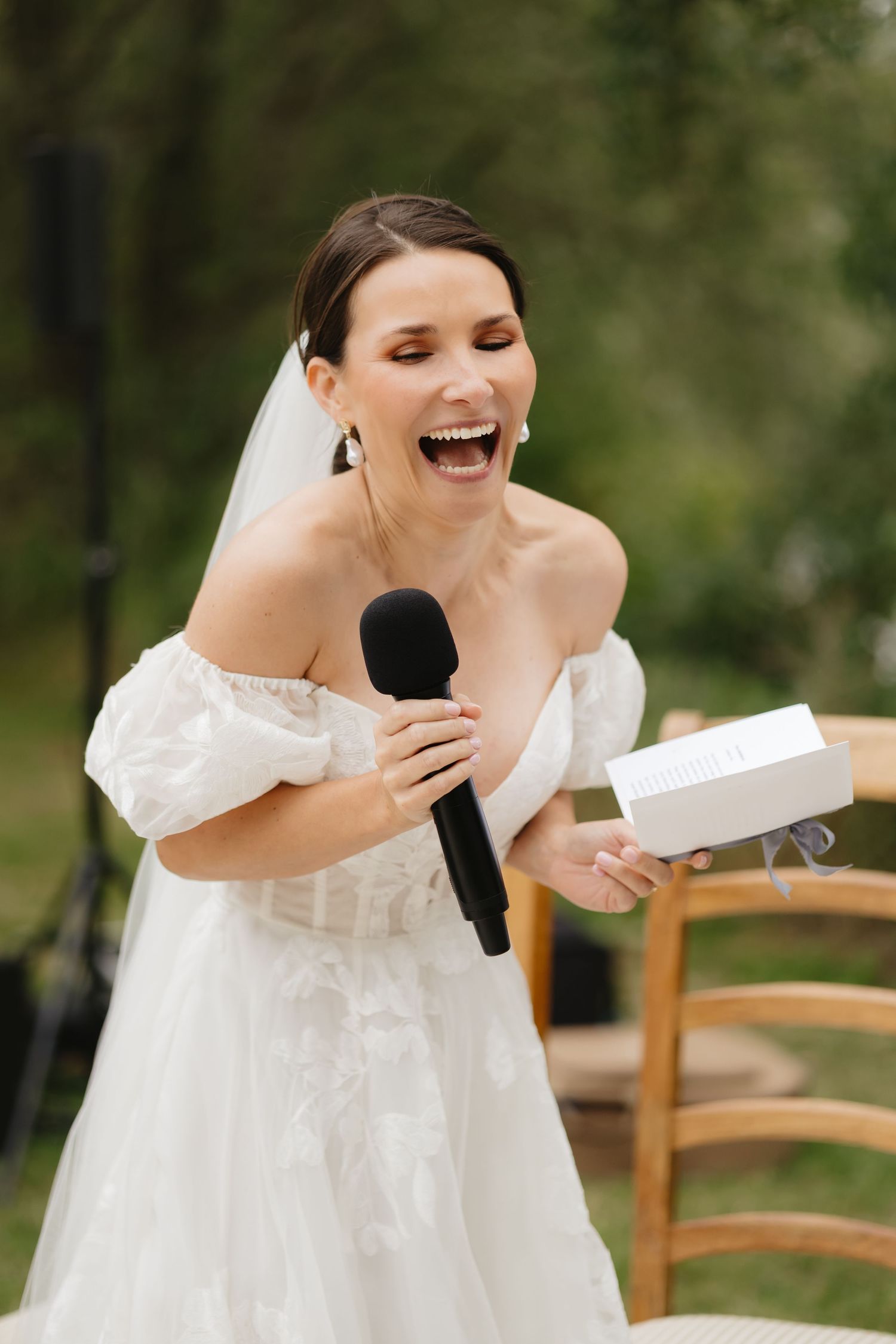 Bride in white dress laughing and covering her face during outdoor ceremony.
