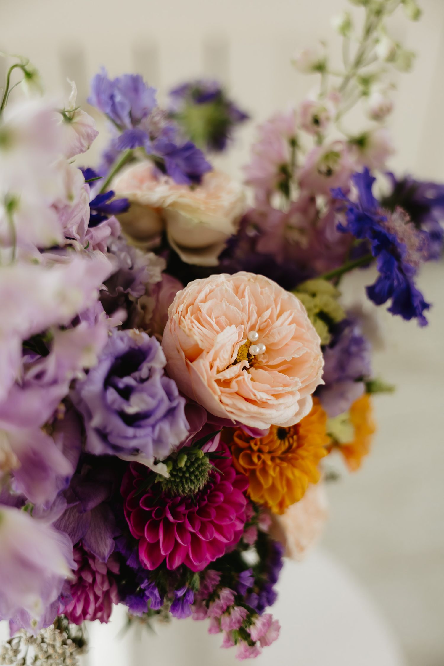Close-up of floral arrangement with peach roses, purple lisianthus, and pink dahlias against a light background.