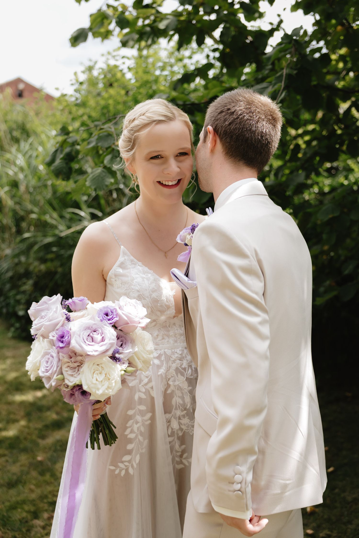 A happy couple shares a moment in their wedding attire, with the bride holding a purple and white bouquet in a garden setting.