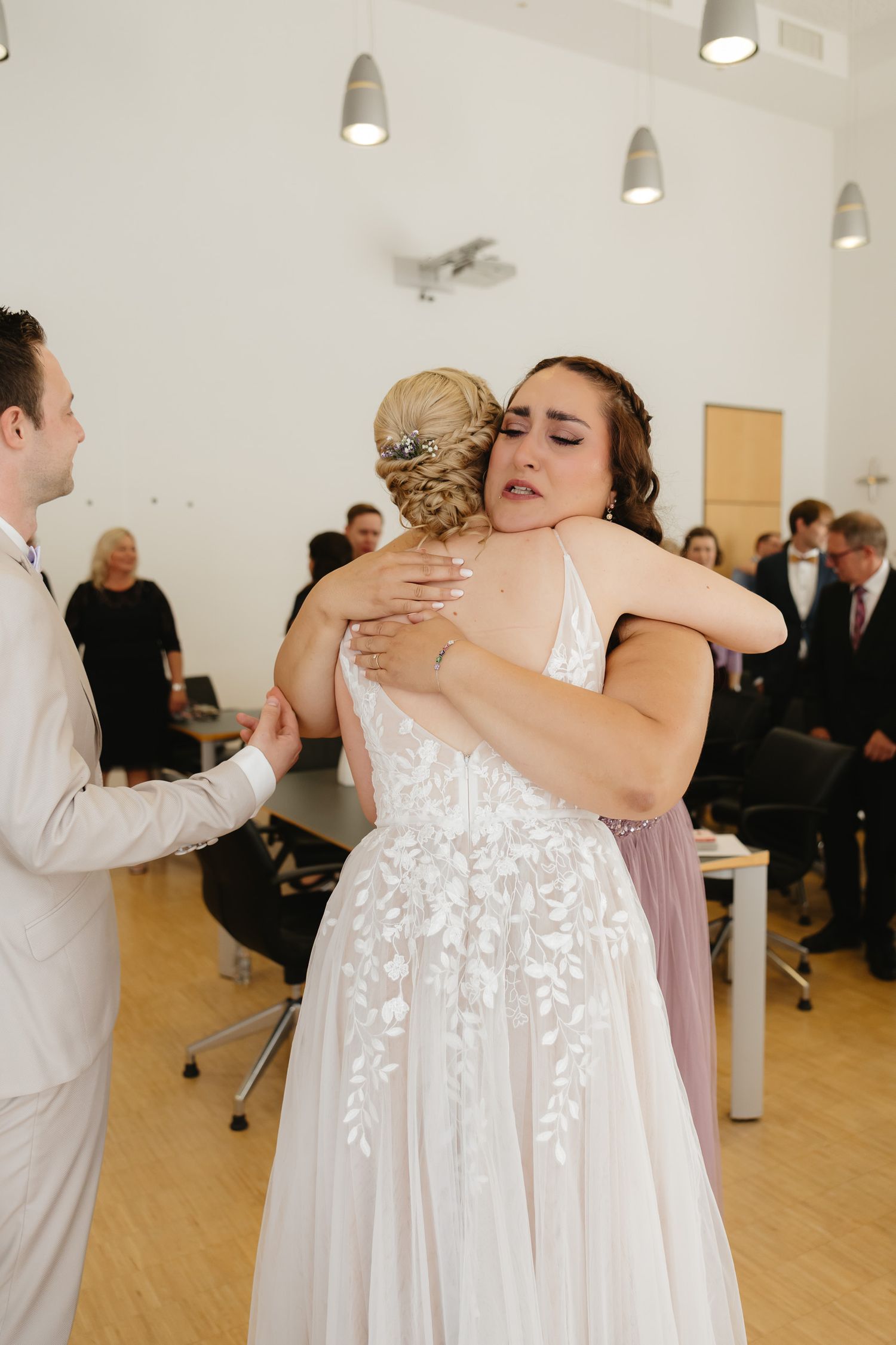 Bridesmaids in flowing white dresses embrace emotionally during a wedding ceremony in a bright indoor venue.