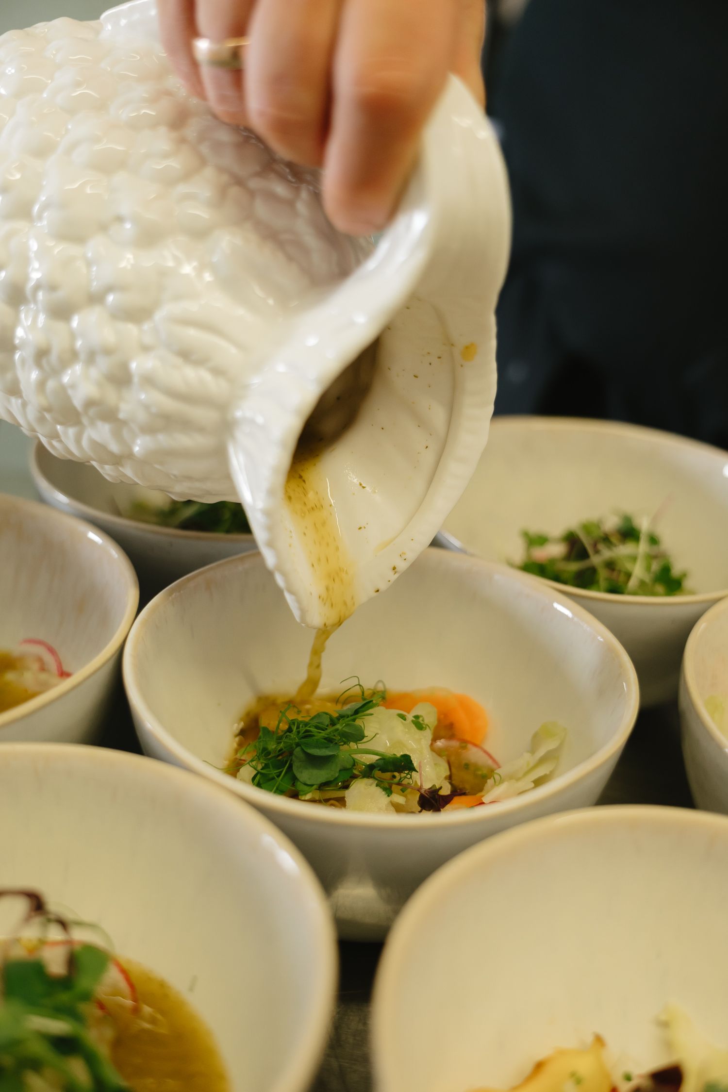 A sequence of hands pouring liquid egg yolks into small white bowls containing vegetables and garnishes.