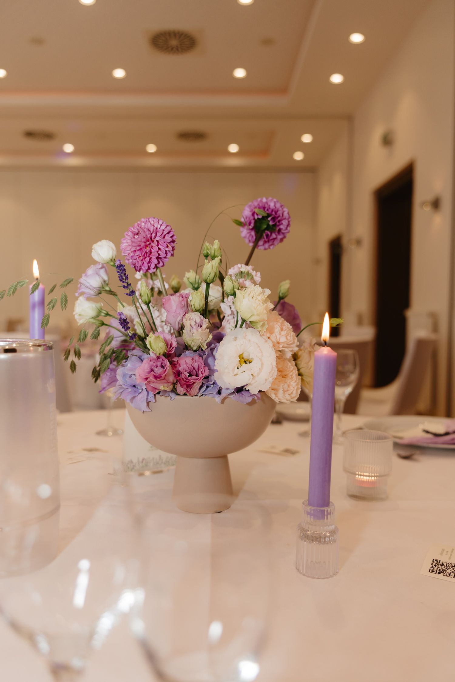 Elegant table setting with purple candles and pastel floral centerpieces in white bowls at an indoor reception.