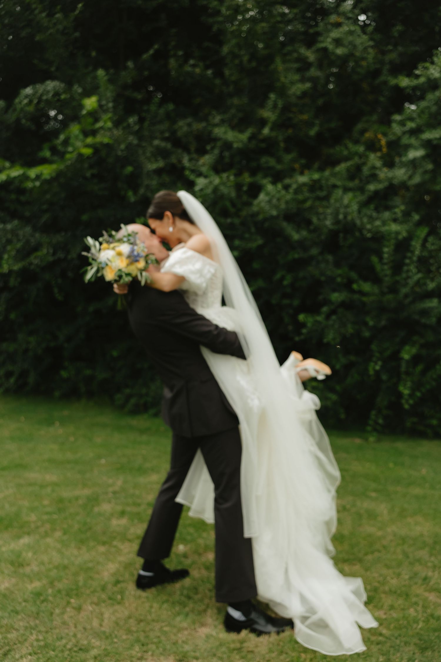 Couple in wedding attire dances gracefully on grass with trees in background.