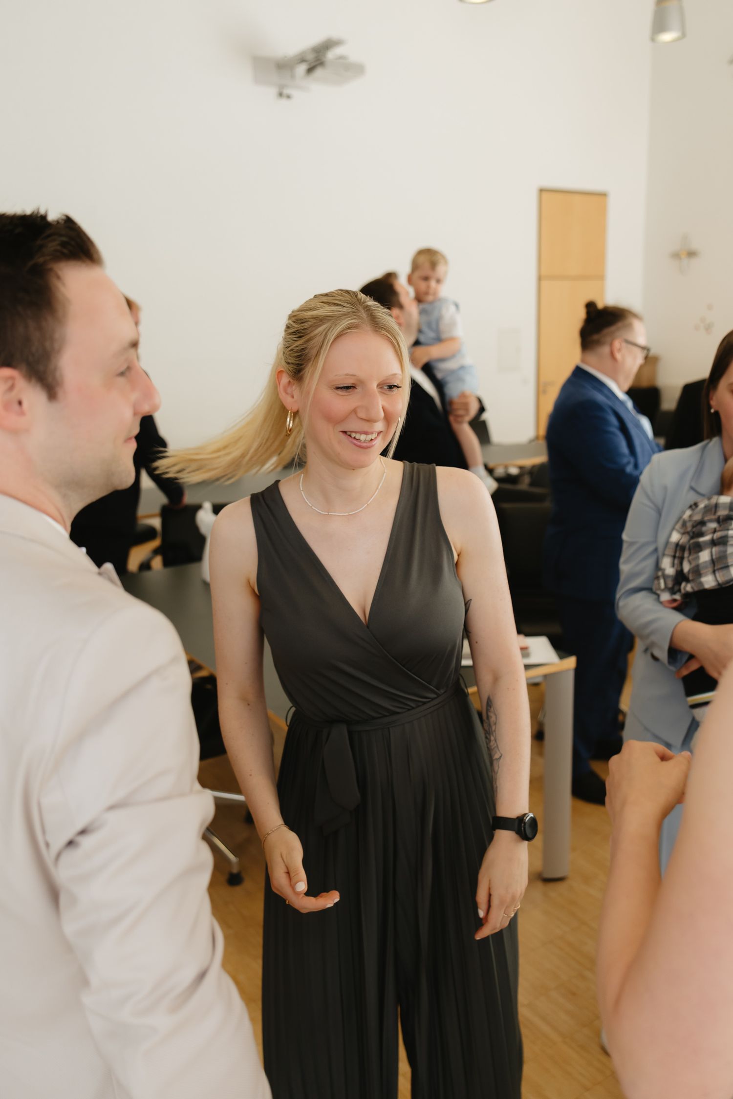 People socializing at an indoor gathering, with someone wearing a black wrap dress in focus at a casual event.