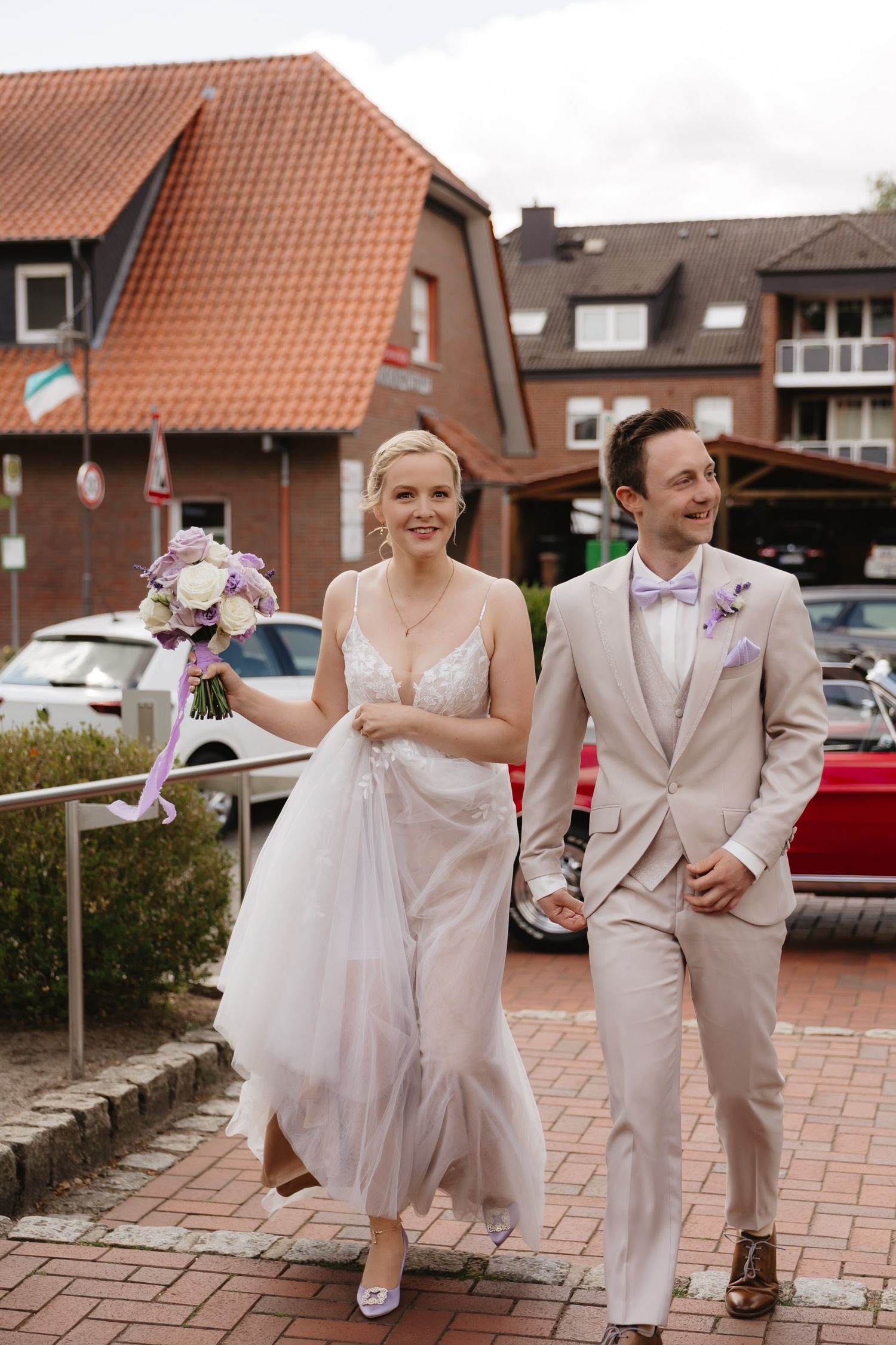 A couple in wedding attire walks together and poses playfully on a brick paved walkway outside brick buildings.