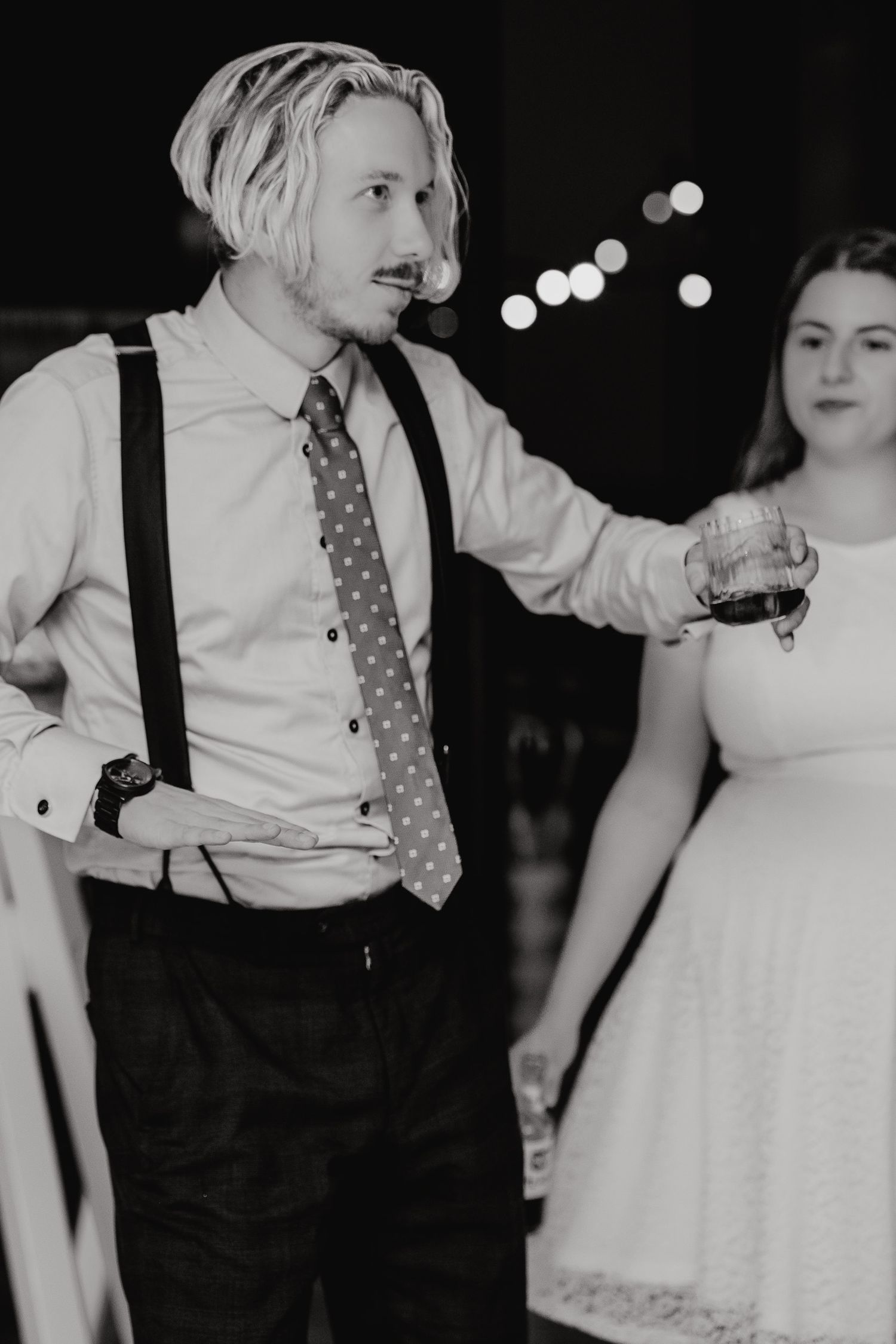 Black and white photo of a wedding reception with a person in suspenders and tie holding a drink next to someone in a white dress.