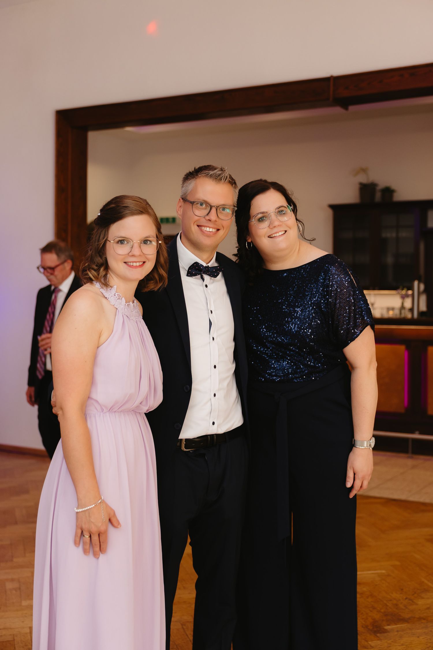 Formal event attendees pose together in evening attire with women in pink and navy gowns and men in black tuxedos.