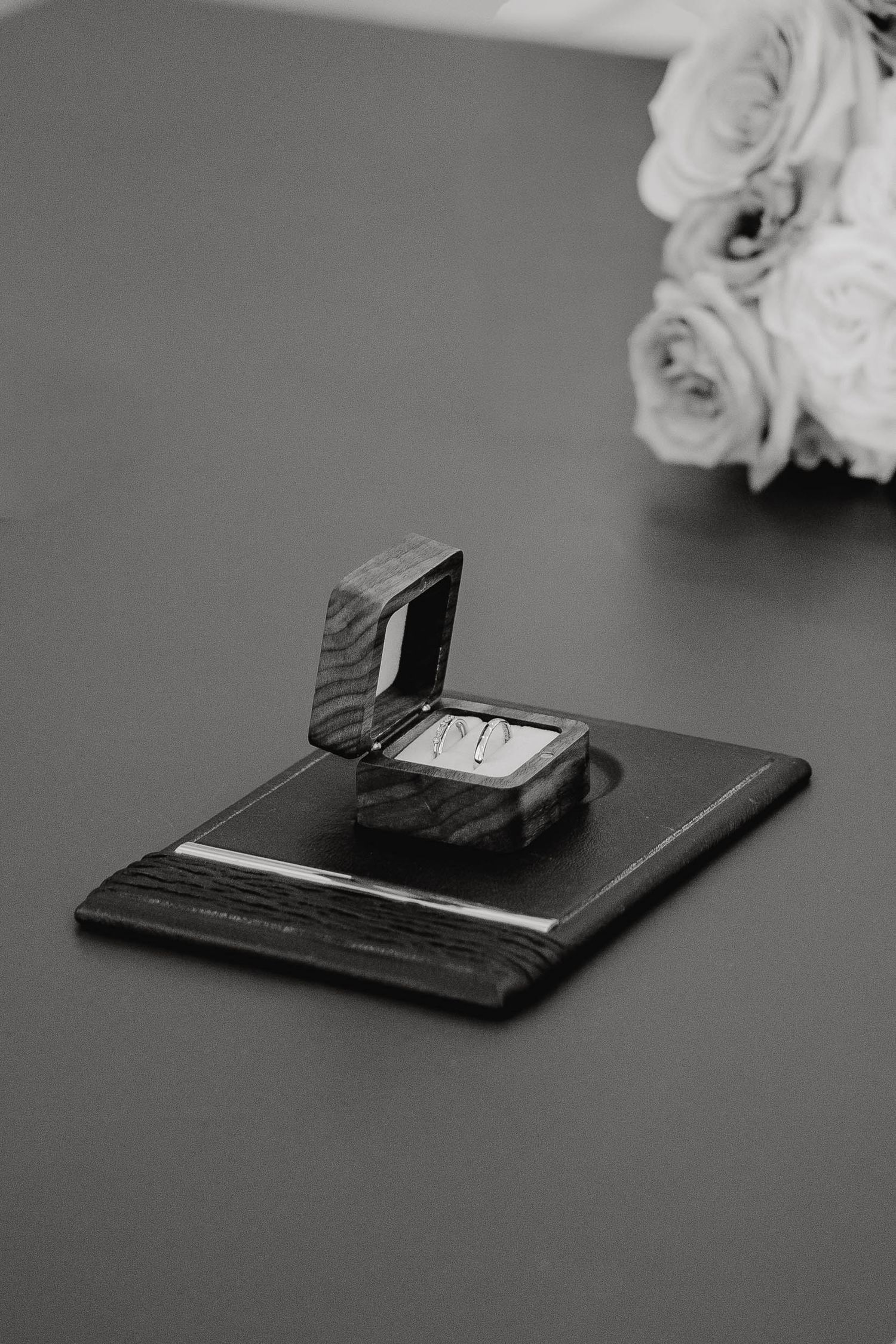 Black and white photograph of a wedding ring box with white roses in soft focus in the background.