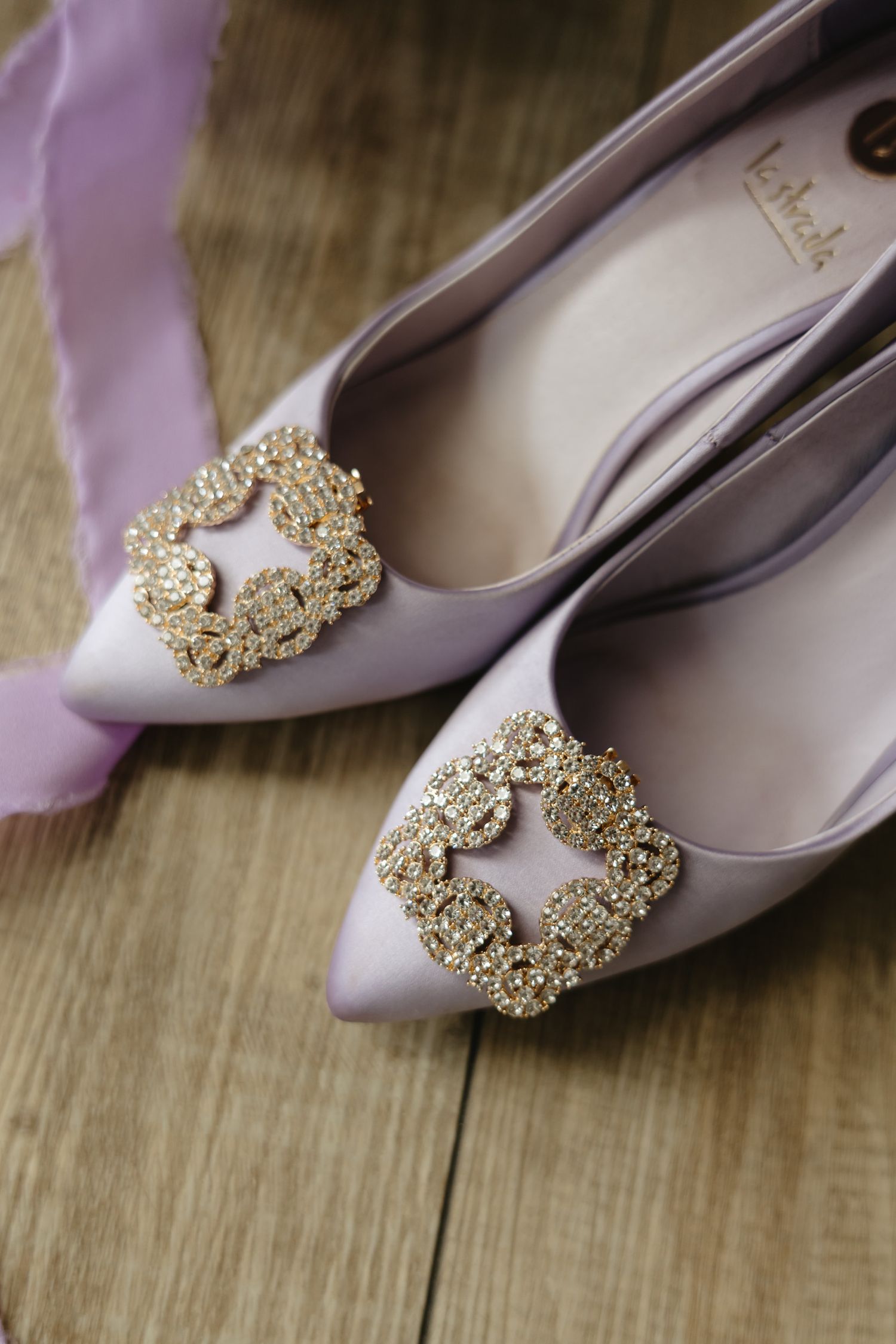 Elegant lavender satin ballet flats adorned with ornate crystal brooches shown from different angles on wooden surface.