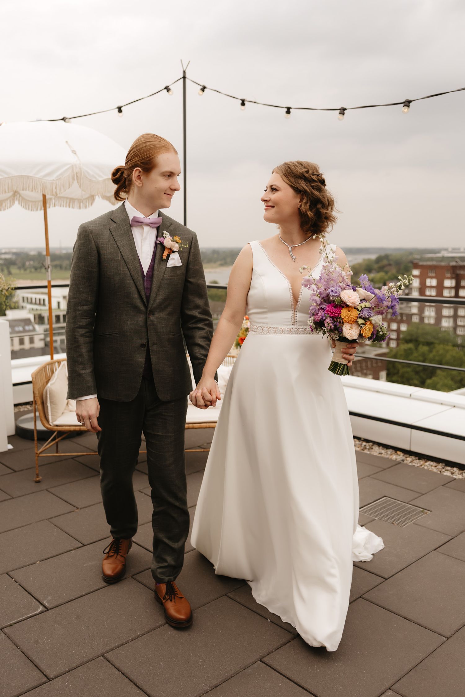 A couple shares a romantic moment on a rooftop terrace decorated with string lights and featuring a colorful bridal bouquet.