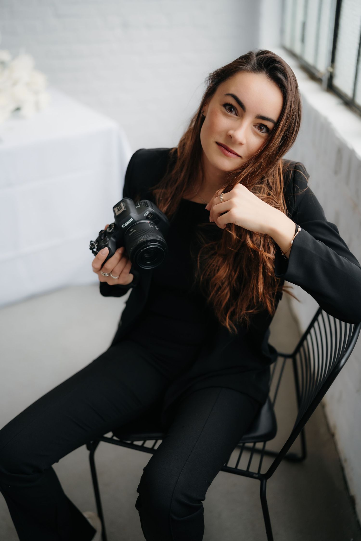 Photographer in black outfit poses with camera in bright studio setting.
