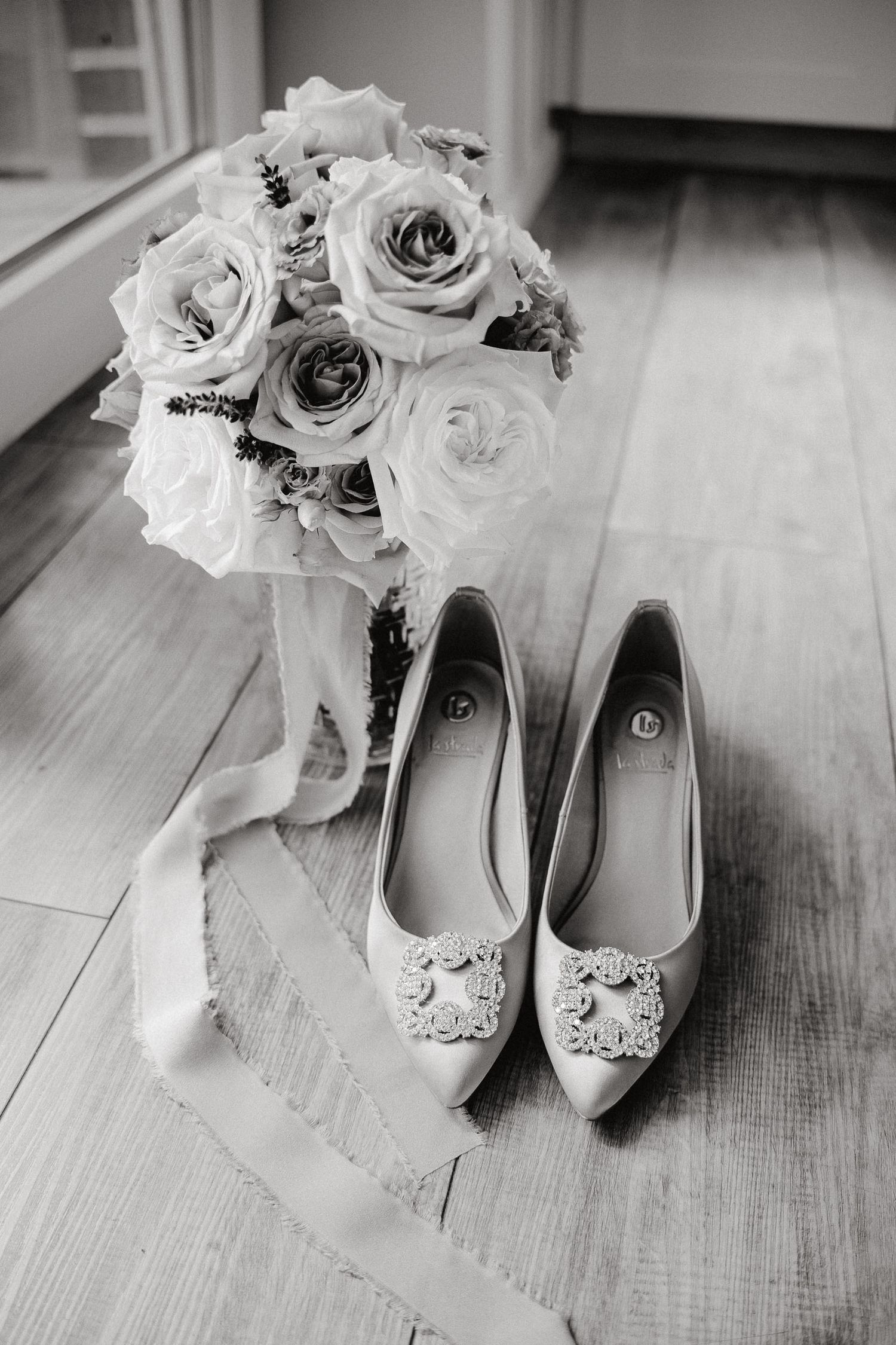 Black and white wedding photos of elegant bridal shoes with floral embellishments paired with roses on wooden floor.