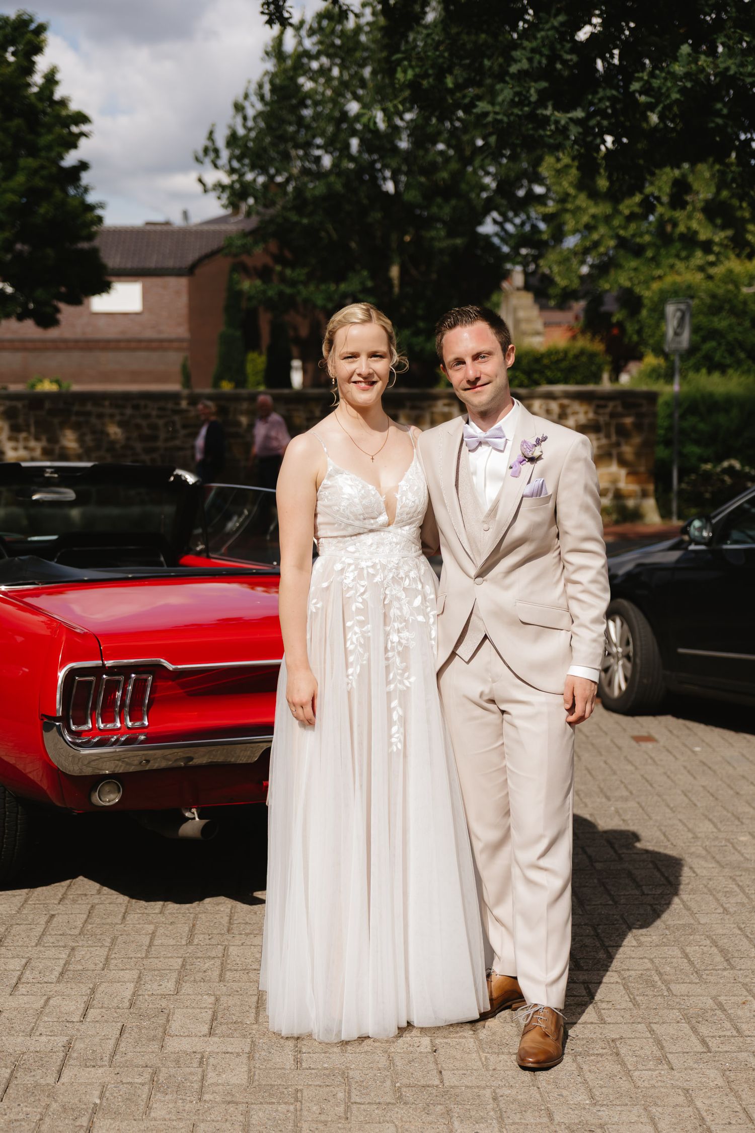 A couple in wedding attire poses together in front of a classic red car on a brick paved driveway.