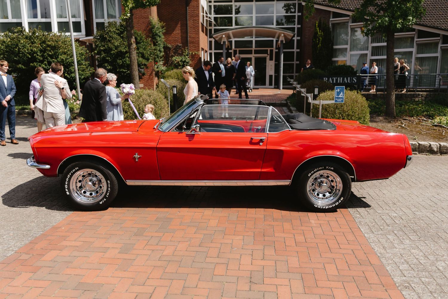 A classic red convertible Mustang drives along a brick paved street near modern buildings.