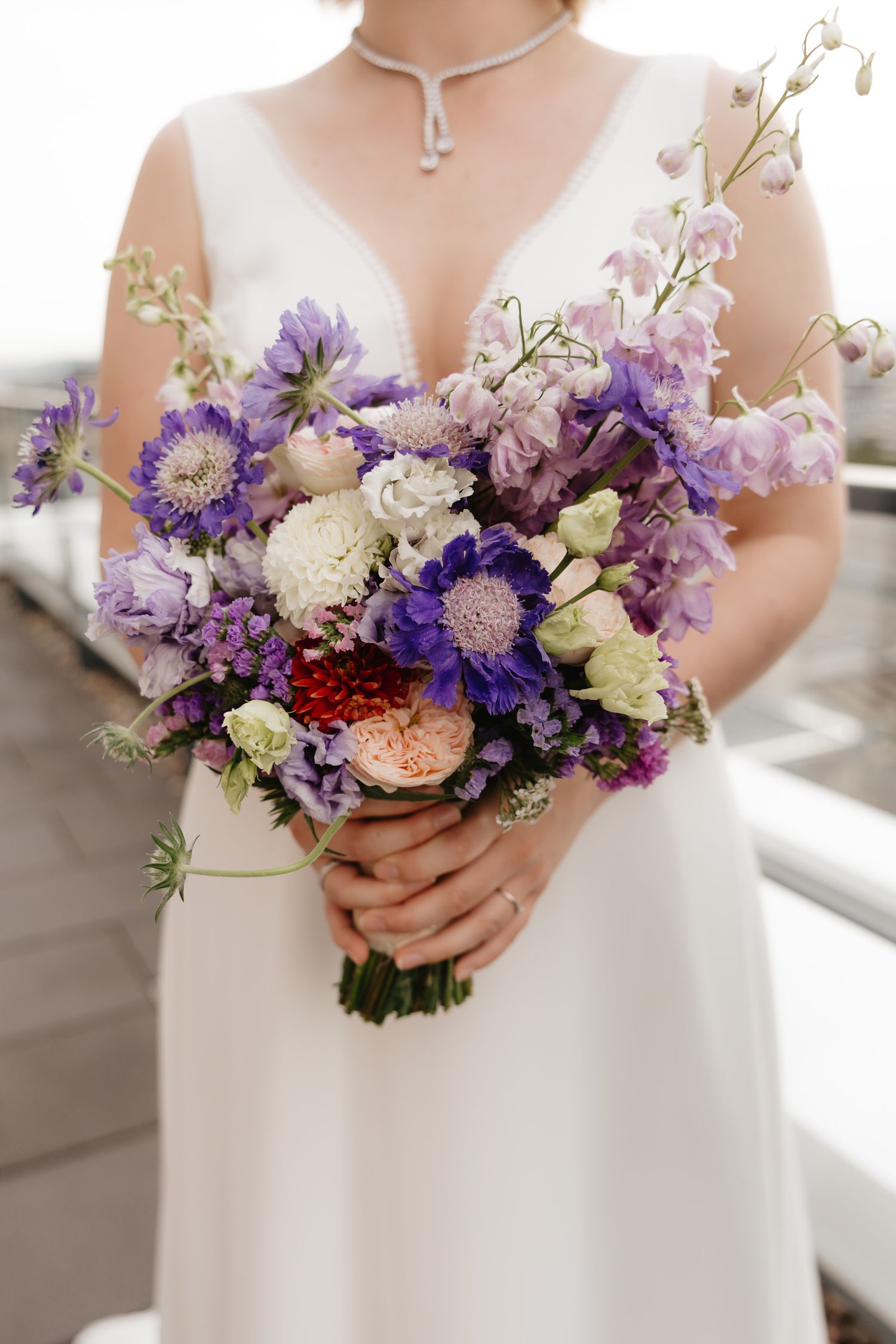 Beautiful purple and white wedding bouquet with mixed flowers held against a white dress in natural light.
