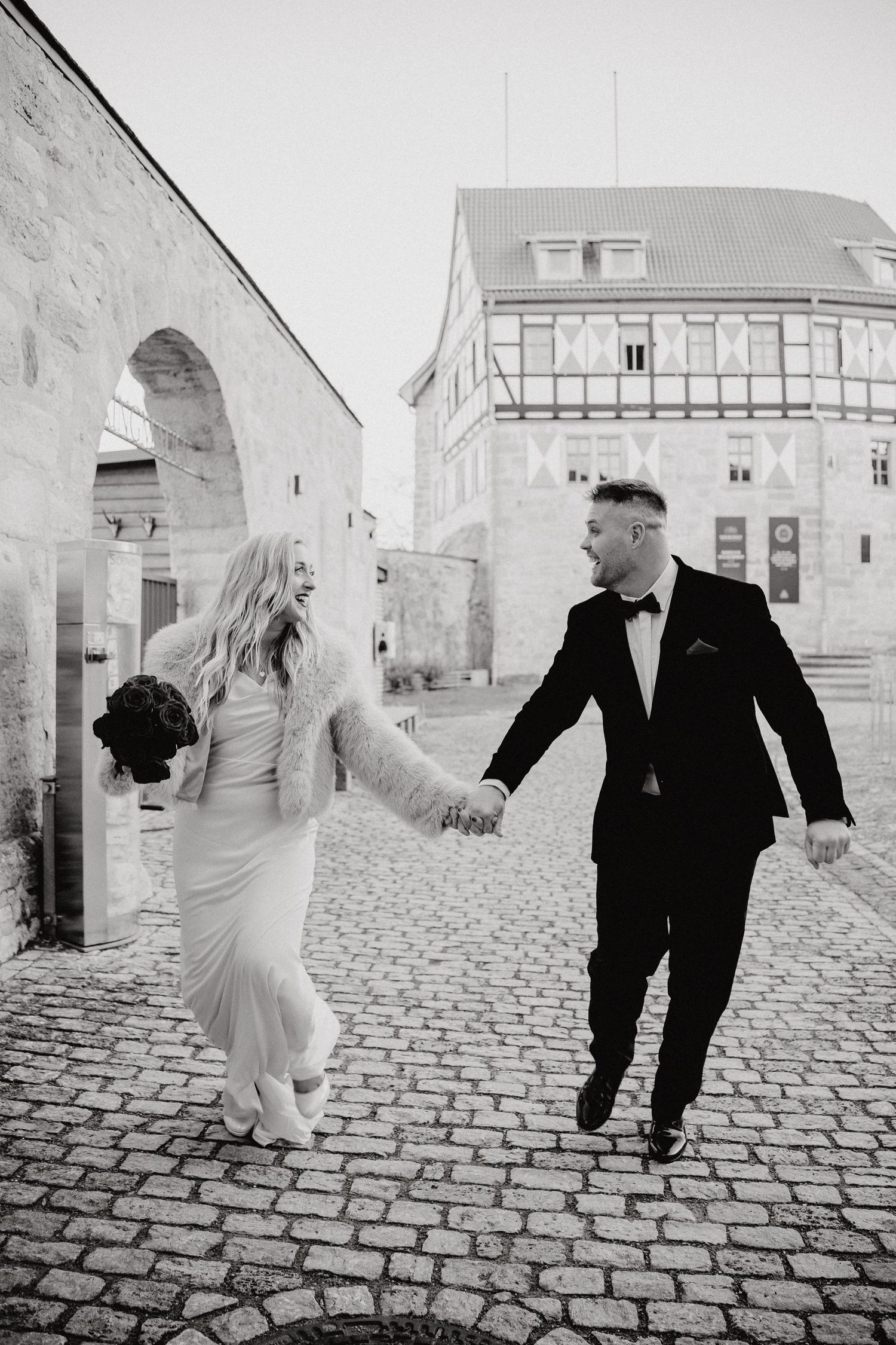 Black and white wedding photos show playful couple dancing on cobblestone street.