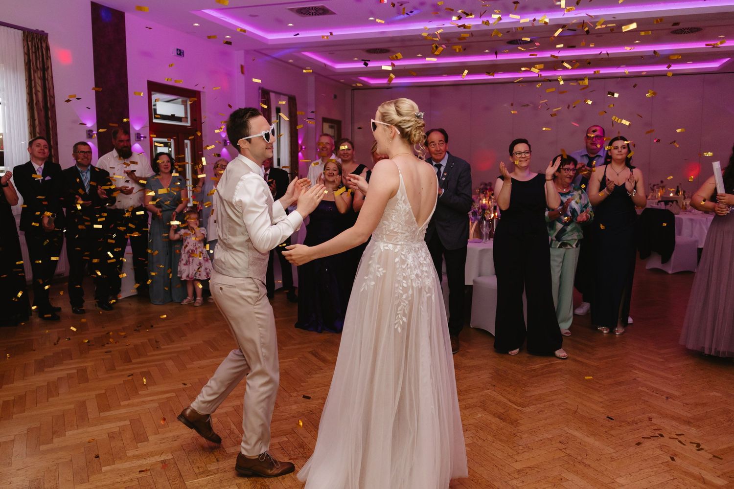 Wedding guests celebrate on a dance floor under pink lighting while confetti falls during a reception celebration.