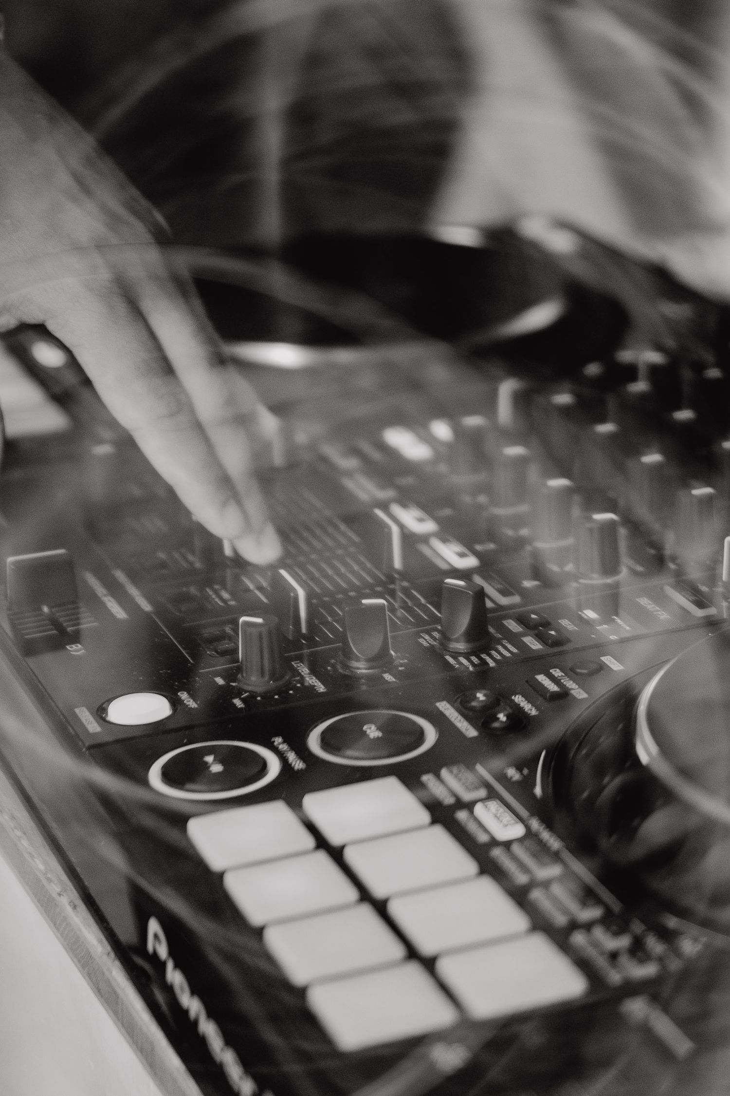 Close-up black and white shot of DJ mixer with knobs, faders and drum pads being operated by a hand.