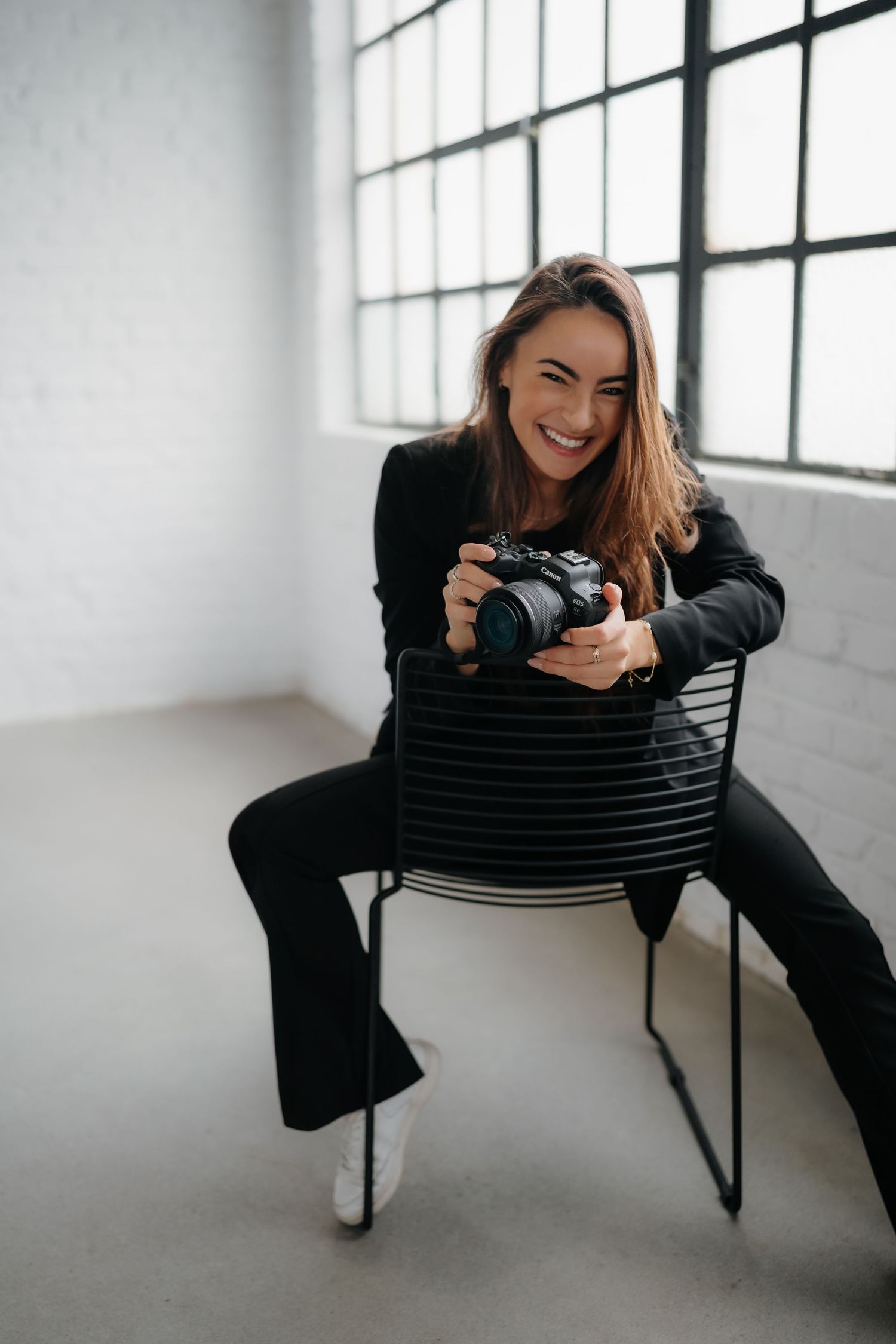 Playful photographer poses with camera on modern chair in bright studio setting.