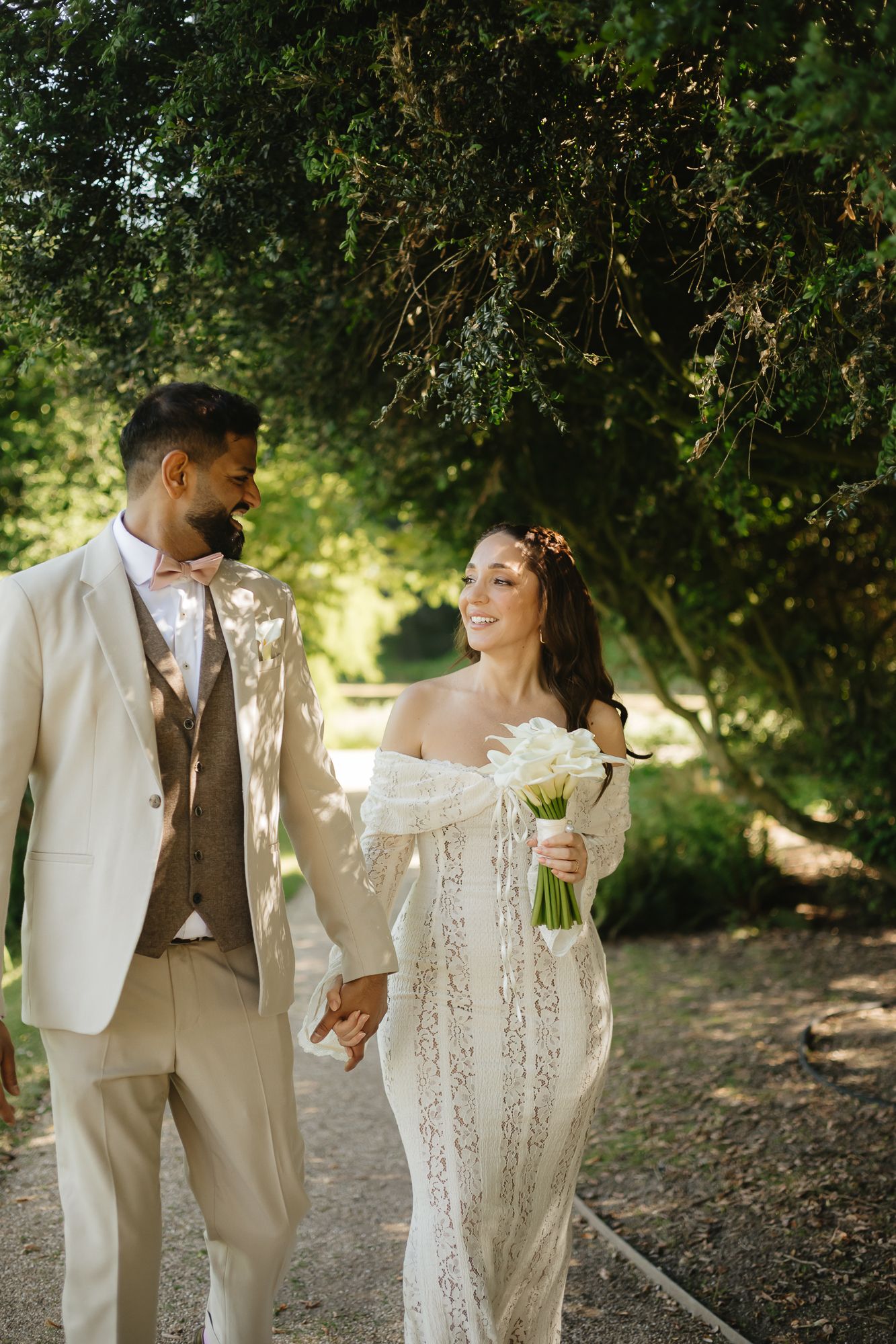 Newlyweds in light-colored attire walking together on a sunlit garden path.