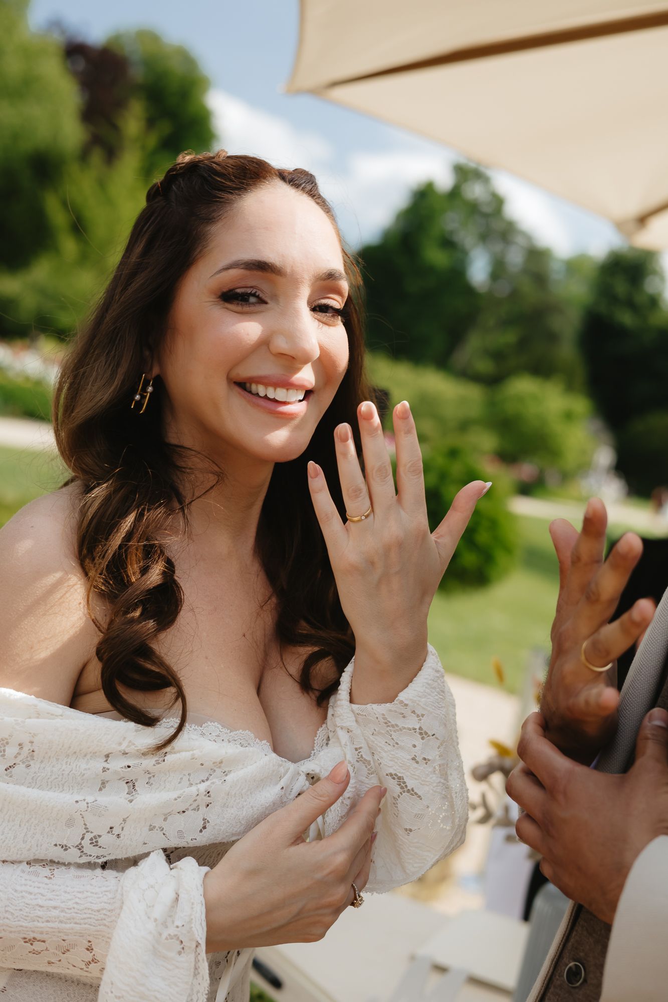 Bride in lace dress shows off new ring during outdoor garden wedding celebration.