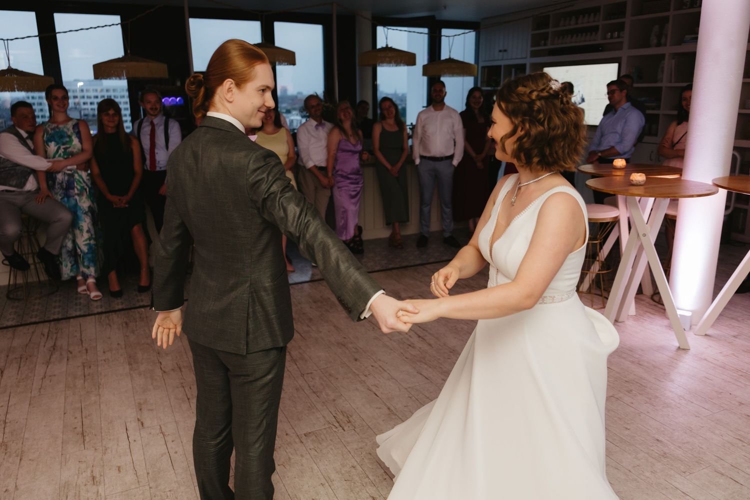 A couple dances together on a wooden floor during their wedding celebration with guests in the background.