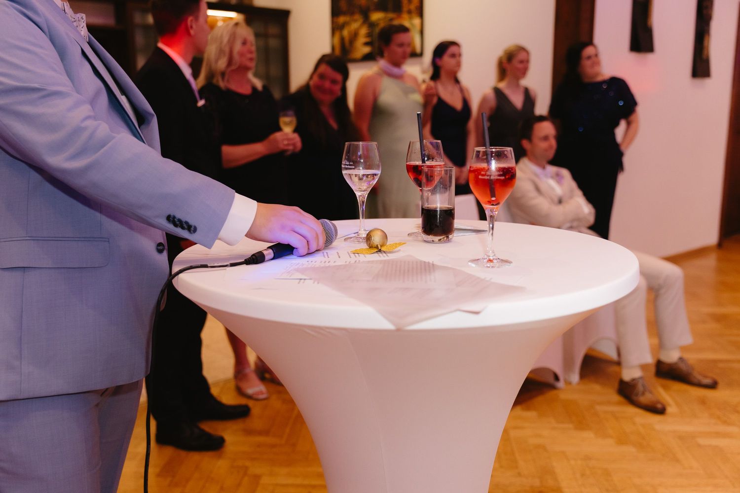 A white cocktail table with wine glasses at an indoor event with guests mingling in the background.