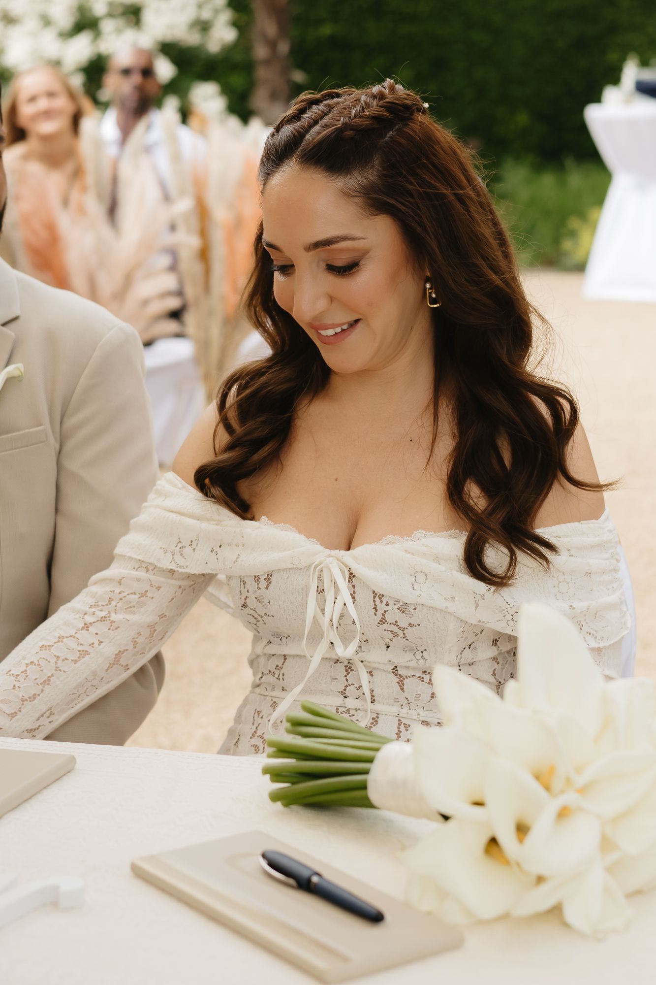 Bride in off-shoulder lace dress signs wedding documents next to white calla lily bouquet at outdoor ceremony.
