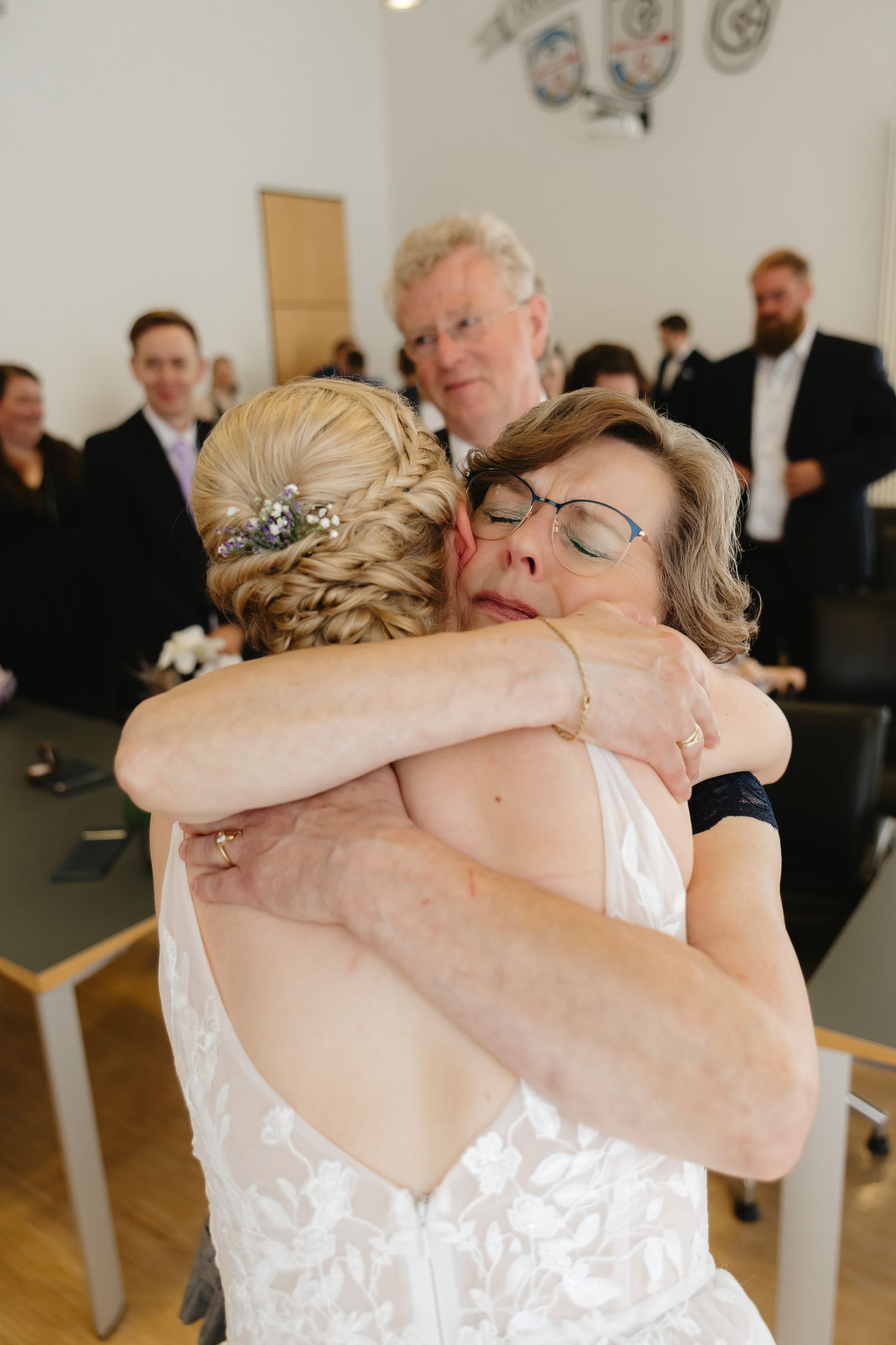 Two people share an emotional embrace during a wedding ceremony in a church with white pews visible in the background.