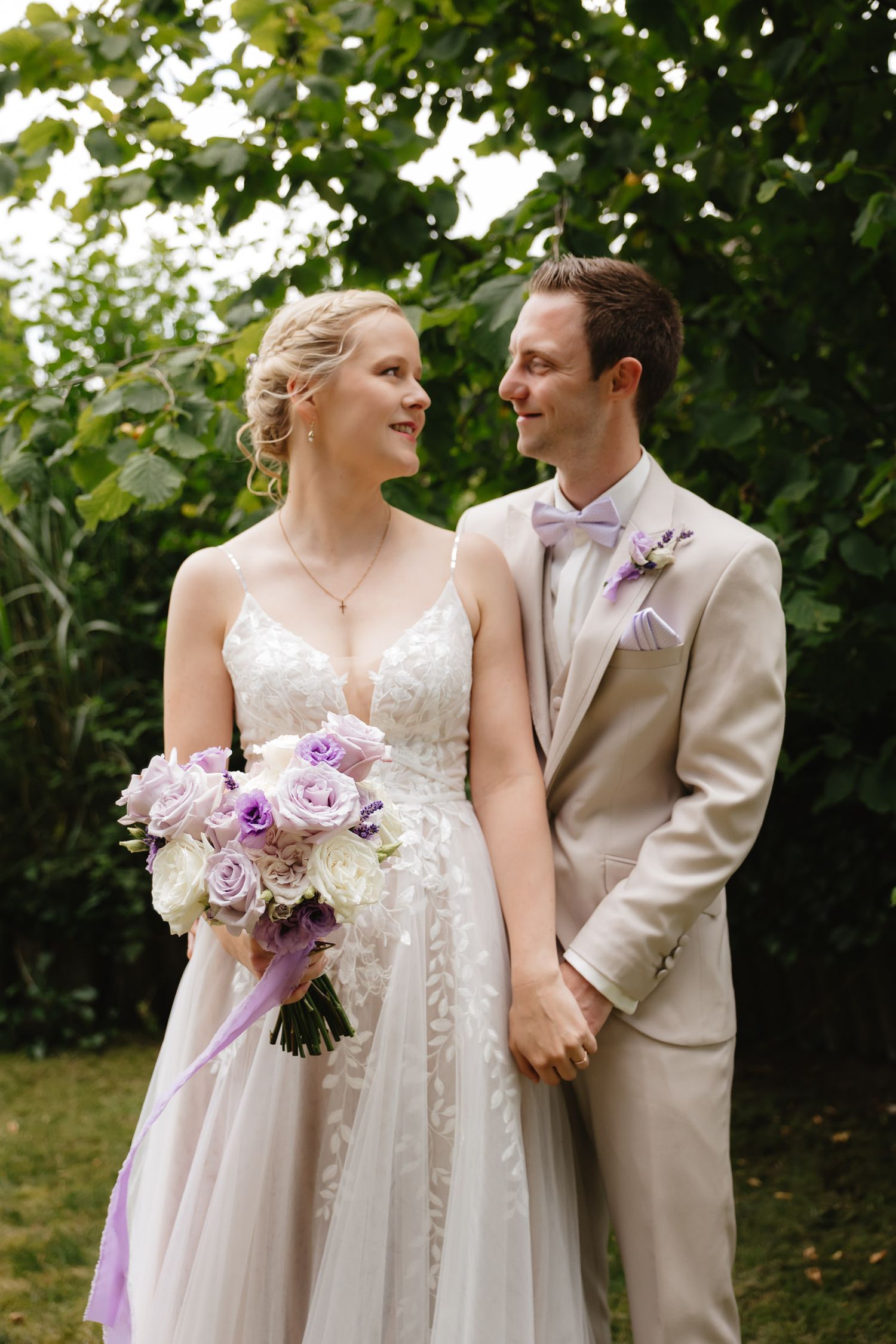 A bride and groom share joyful moments in a garden setting with purple and lavender floral bouquet details.