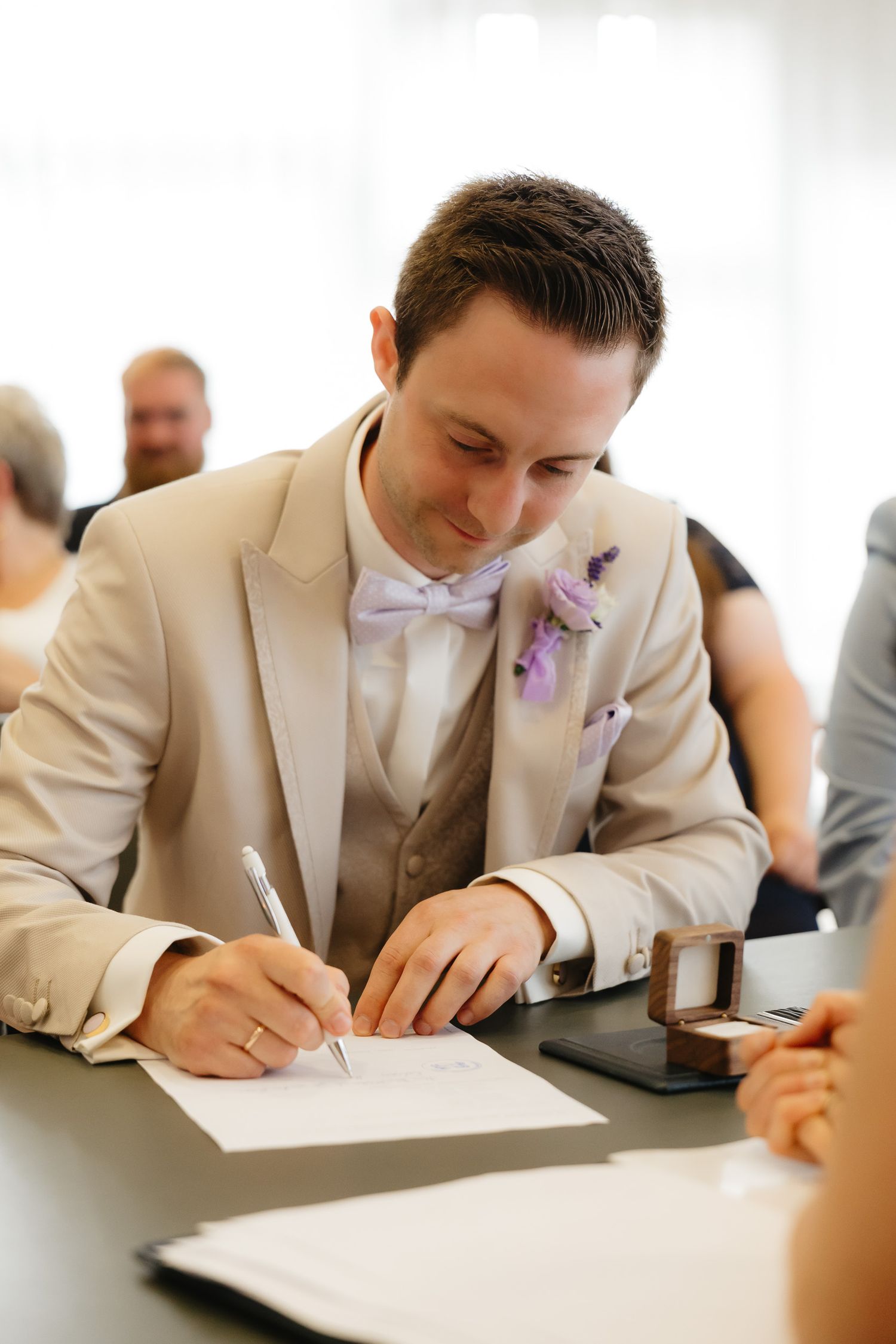 A groom in a beige suit signs marriage documents at a wedding ceremony.