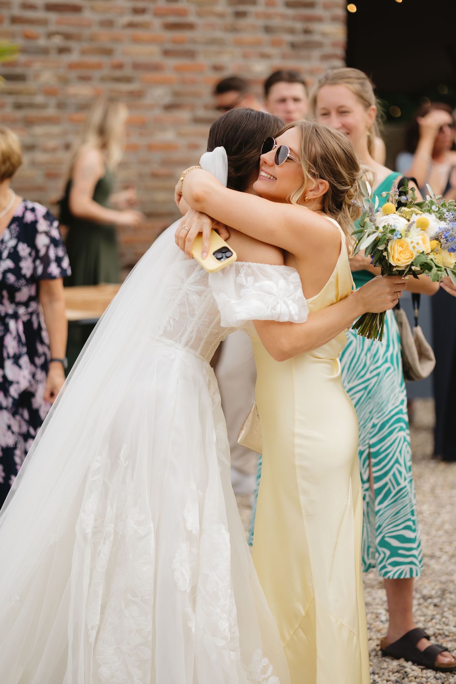 Wedding guests blow bubbles during an outdoor celebration near a brick wall.