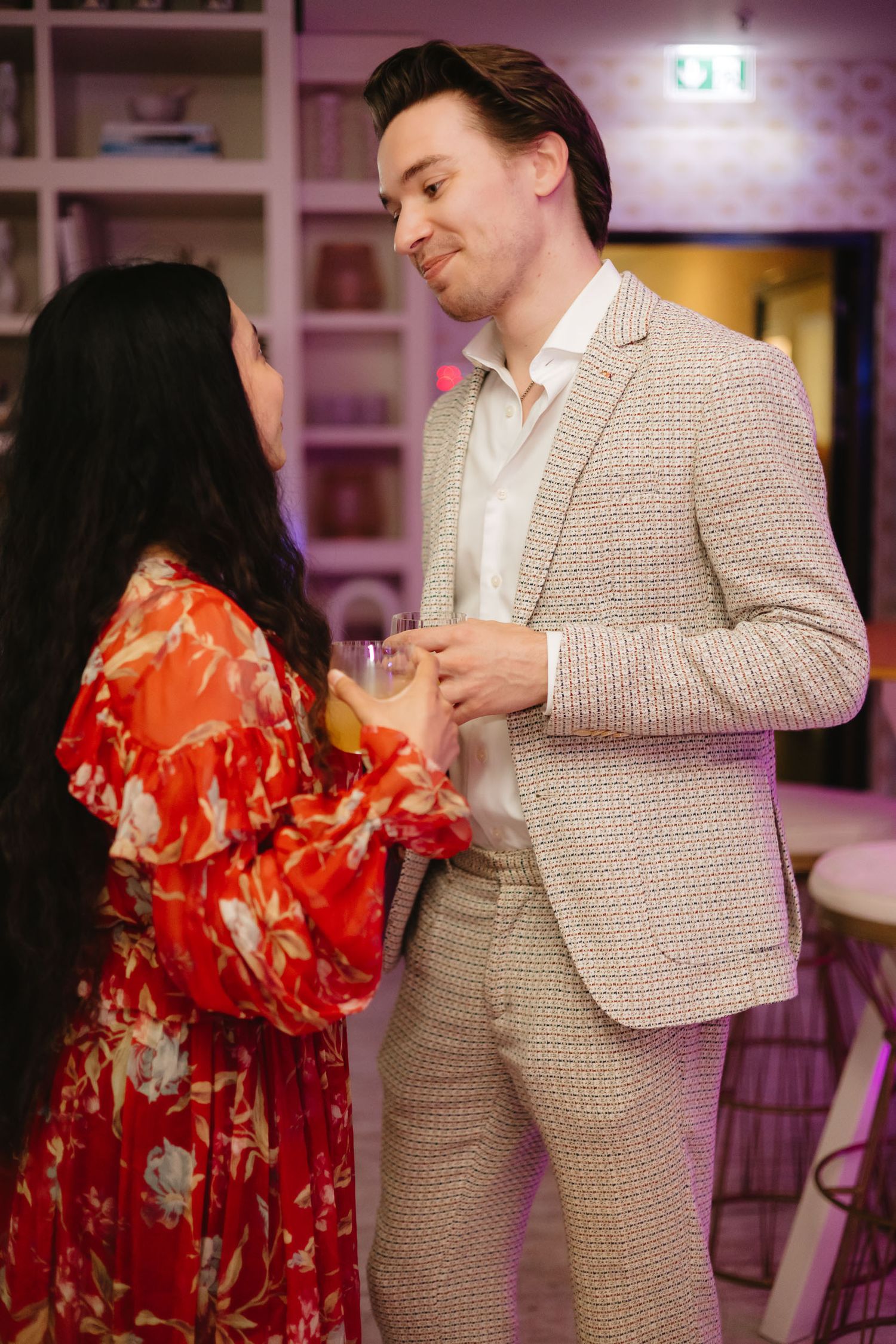 A couple dances together at an event, with one wearing a red floral dress and the other in a beige checkered suit.