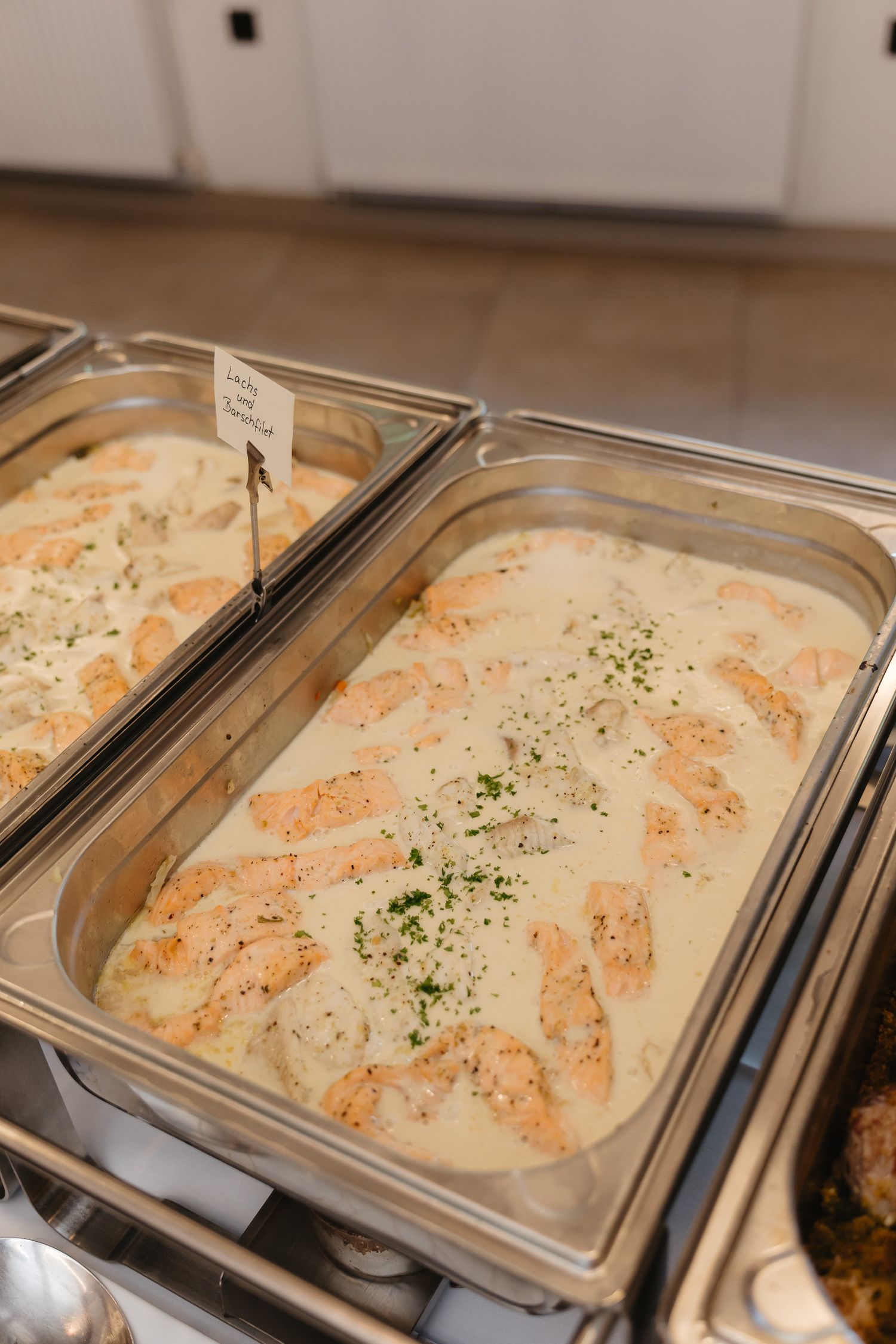 Close-up view of creamy white sauce with herbs garnishing food in metal serving trays at a buffet line.