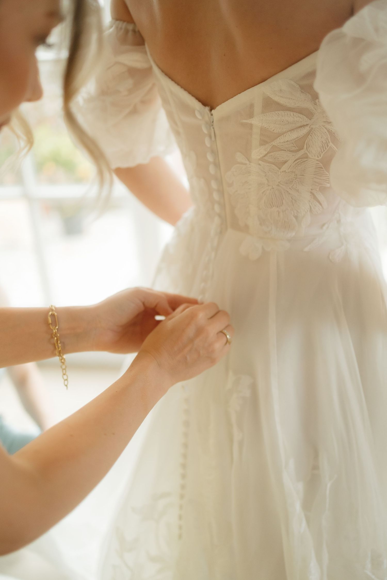Delicate details of a white wedding dress being adjusted in soft natural light.