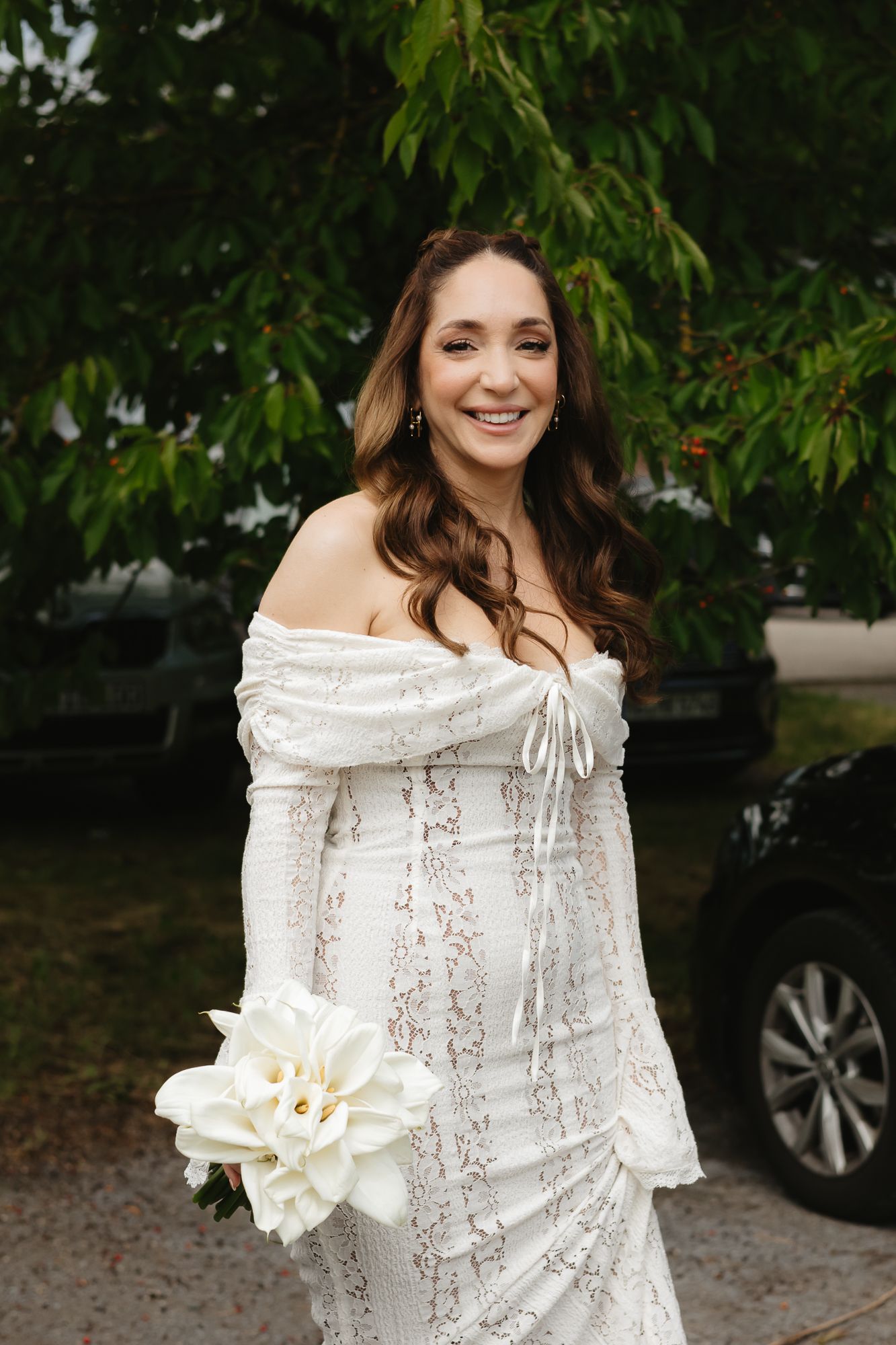 A white lace dress with off-shoulder detail and a white flower bouquet against a garden background.