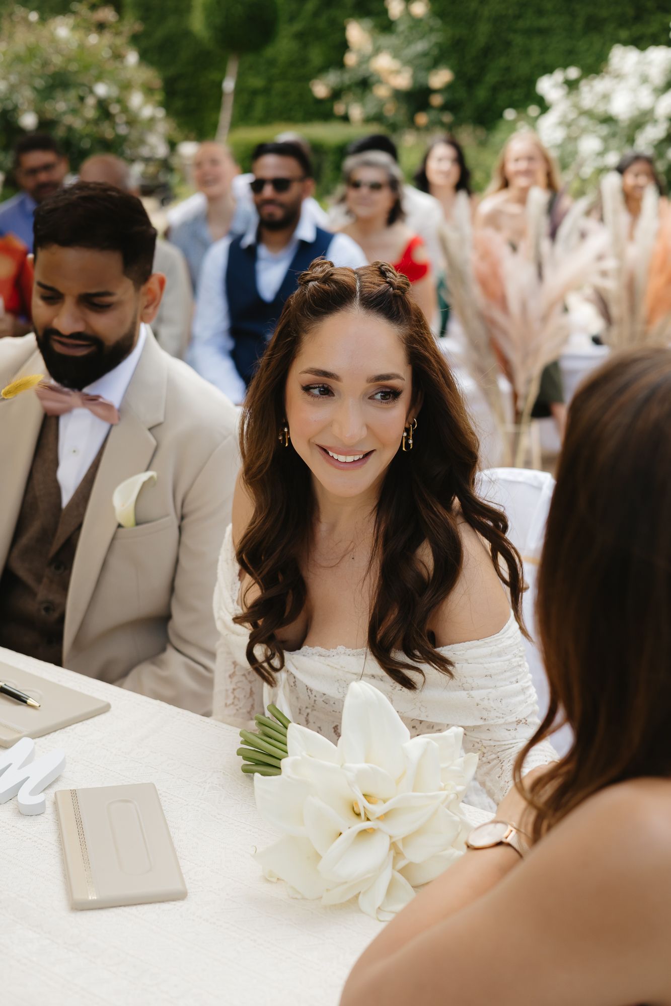 Wedding guests gather around a table during an outdoor celebration with white floral decorations.