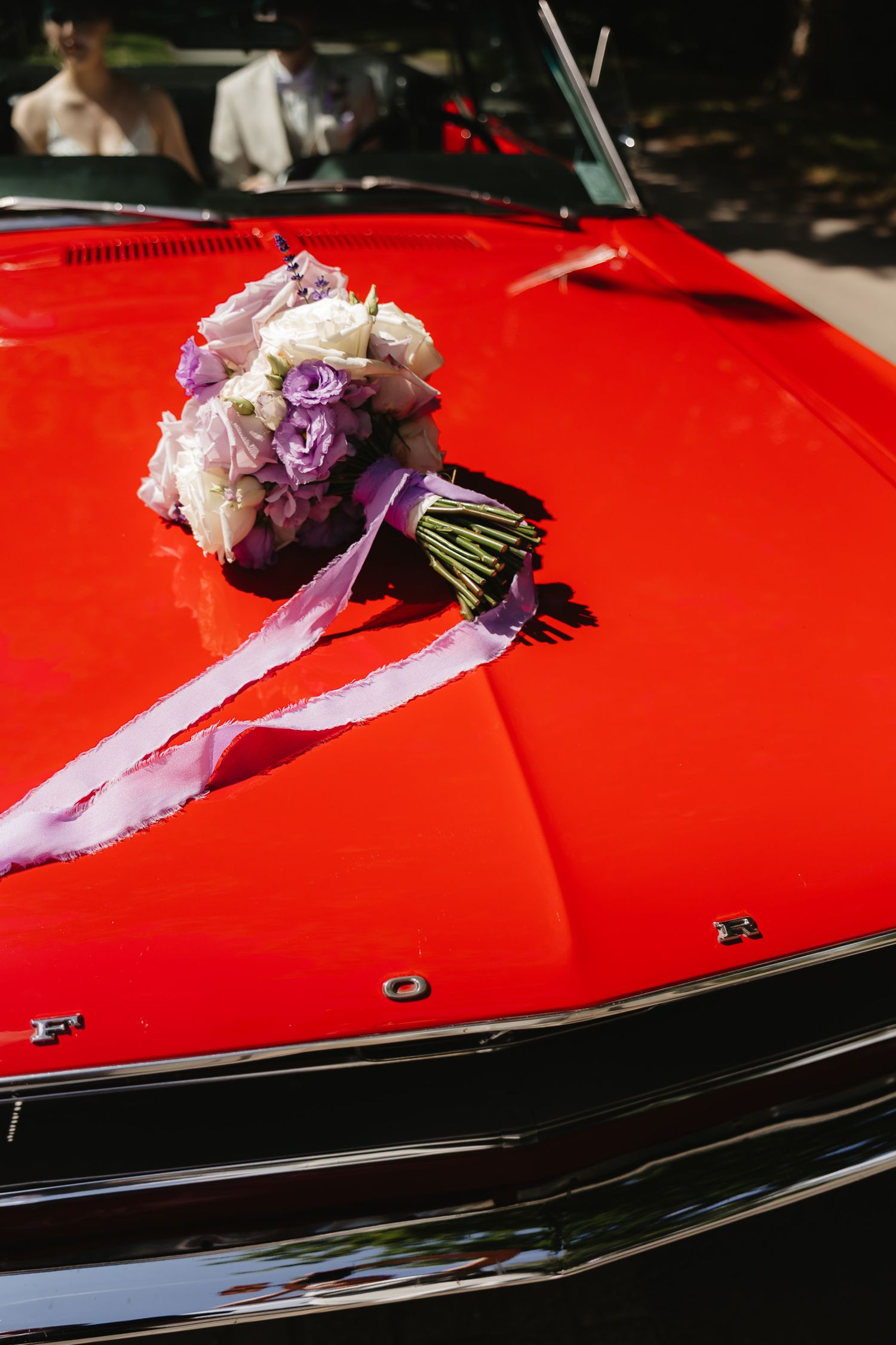 A small floral bouquet with purple ribbons rests on the hood of a shiny red classic car.
