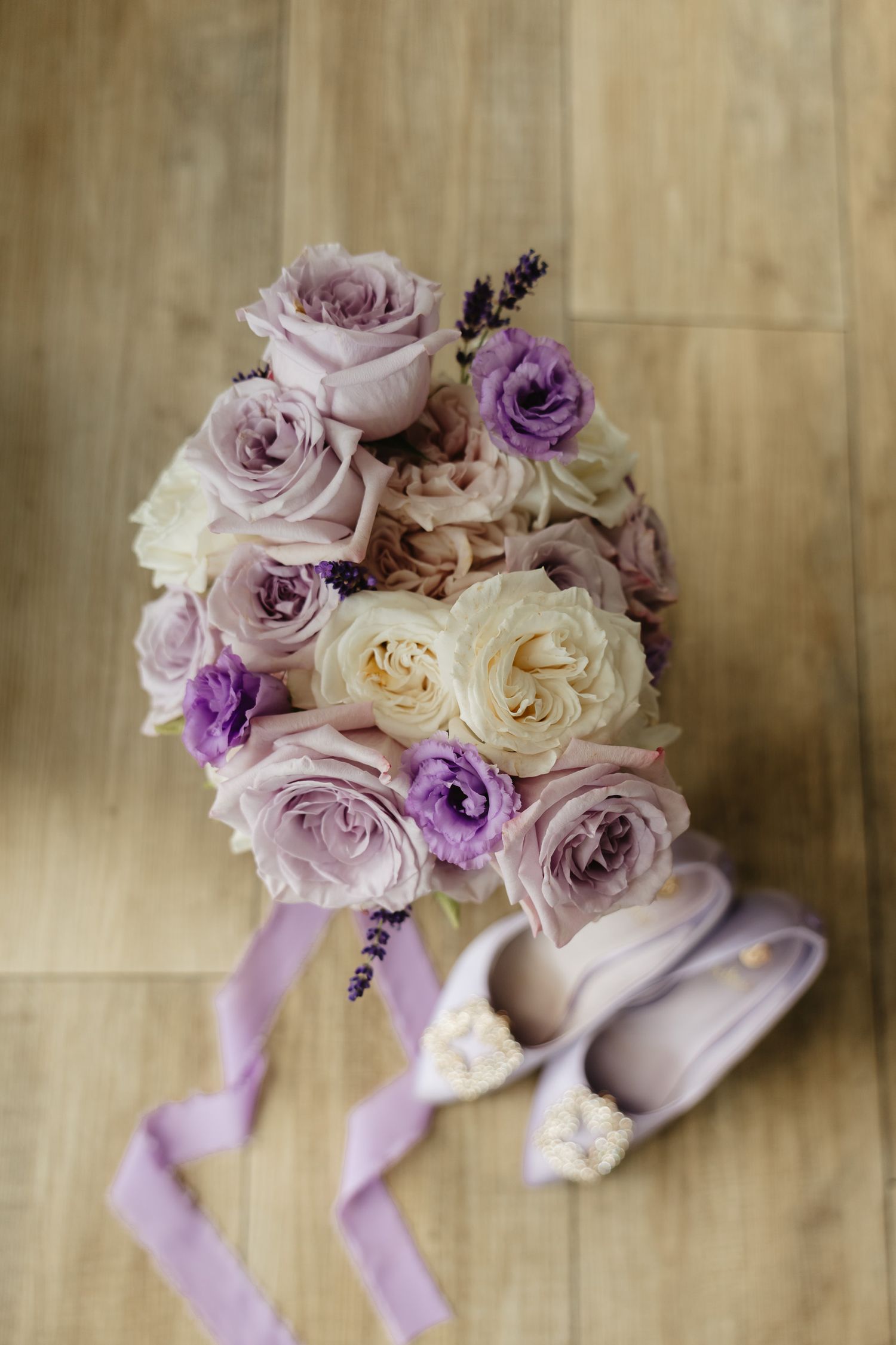 Elegant bridal bouquet with lavender, purple and cream roses arranged with delicate ribbons against a wooden background.