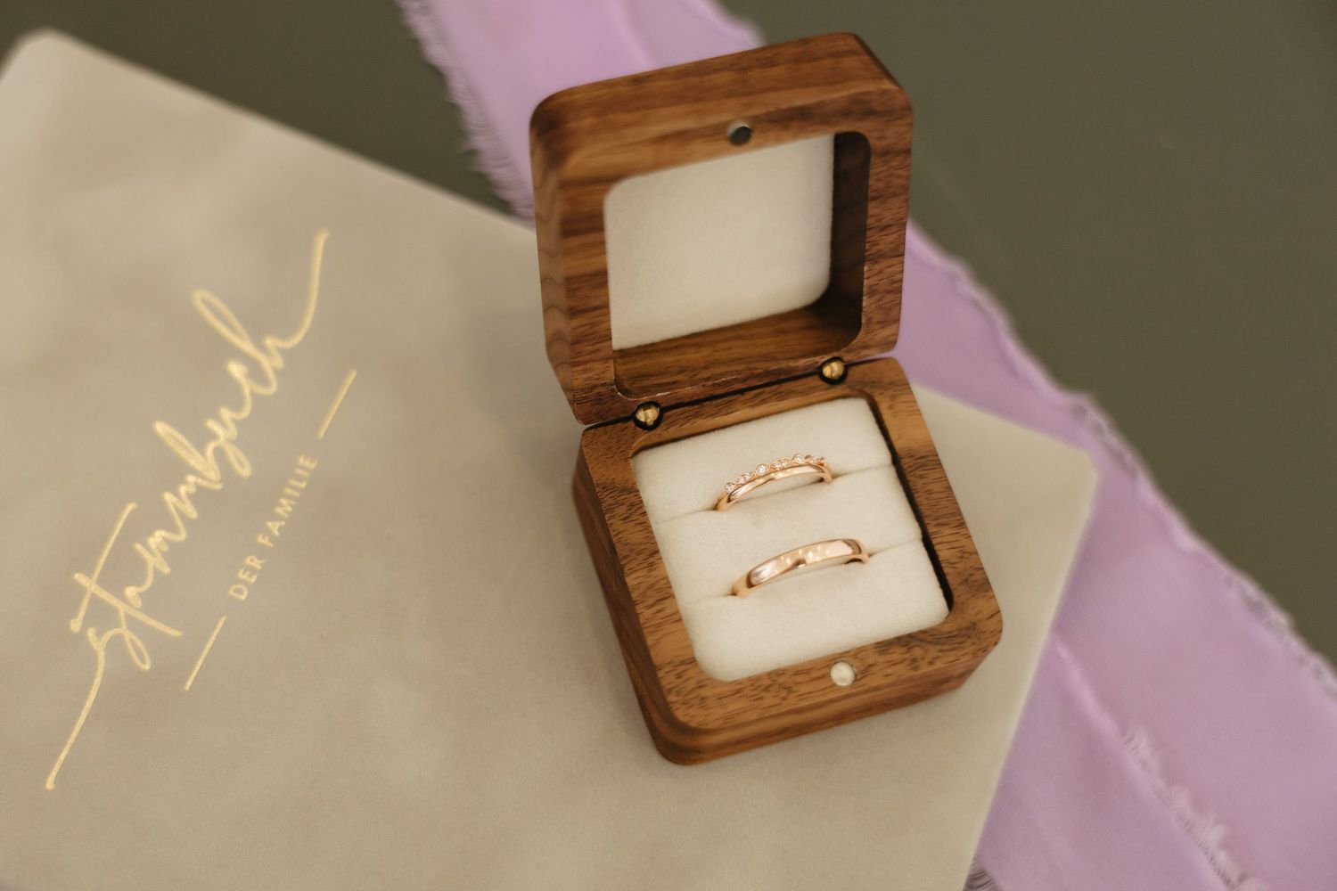 A wooden ring box with gold wedding bands displayed on white cushioning against a purple fabric background.